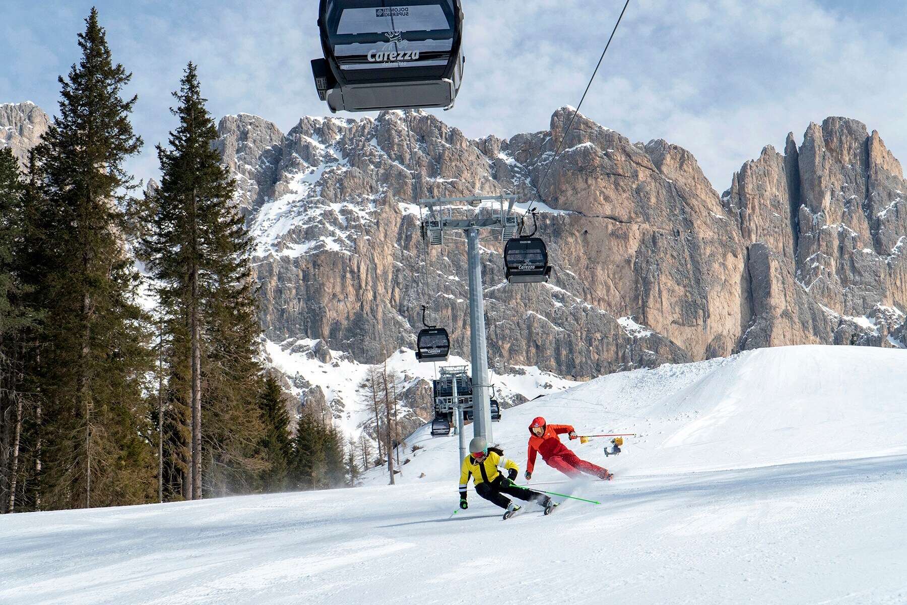 Zwei Skifahrende fahren unter einer Kabinenbahn auf einer Piste vor einem imposanten Bergmassiv