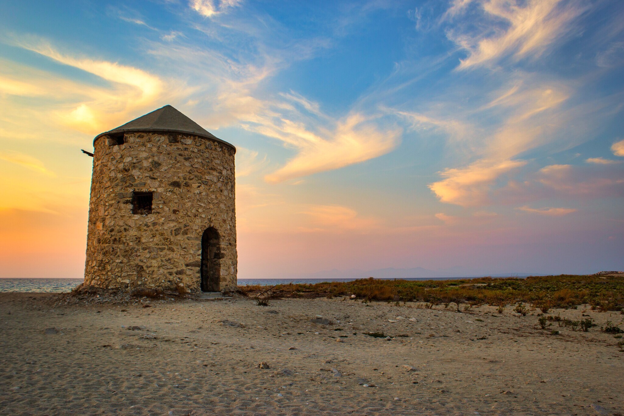 Historische Windmühle aus Stein an einem Strand bei Sonnenuntergang.