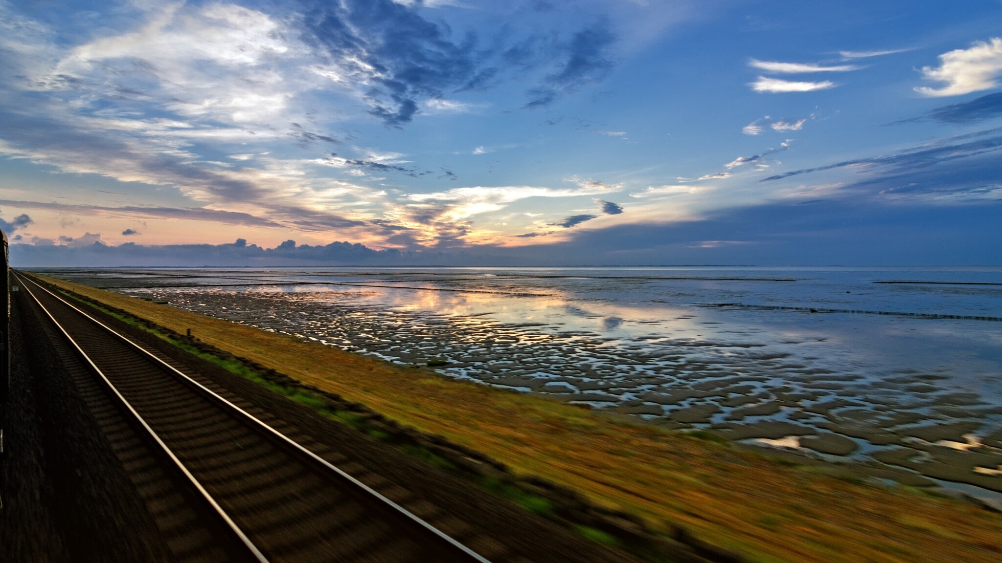 Blick auf den Hindenburgdamm, mit Schienen im Vordergrund und einem weiten Himmel mit Wolken und Sonnenuntergang im Hintergrund. Blick auf den Hindenburgdamm, mit Schienen im Vordergrund und einem weiten Himmel mit Wolken und Sonnenuntergang im Hintergrund.