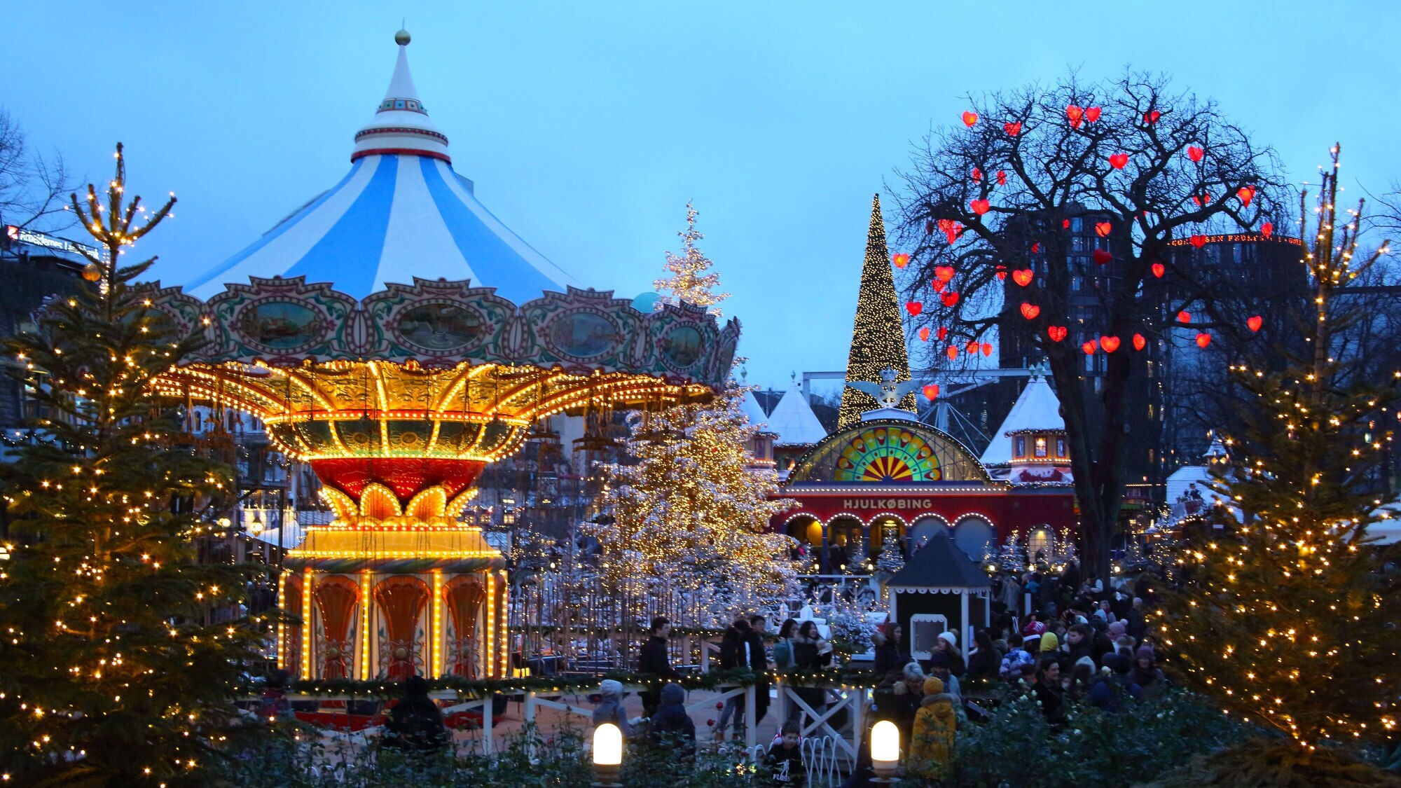Personen in einem Freizeitpark mit historischem Karussell und weihnachtlich beleuchteten Bäumen bei Abenddämmerung. Personen in einem Freizeitpark mit historischem Karussell und weihnachtlich beleuchteten Bäumen bei Abenddämmerung.