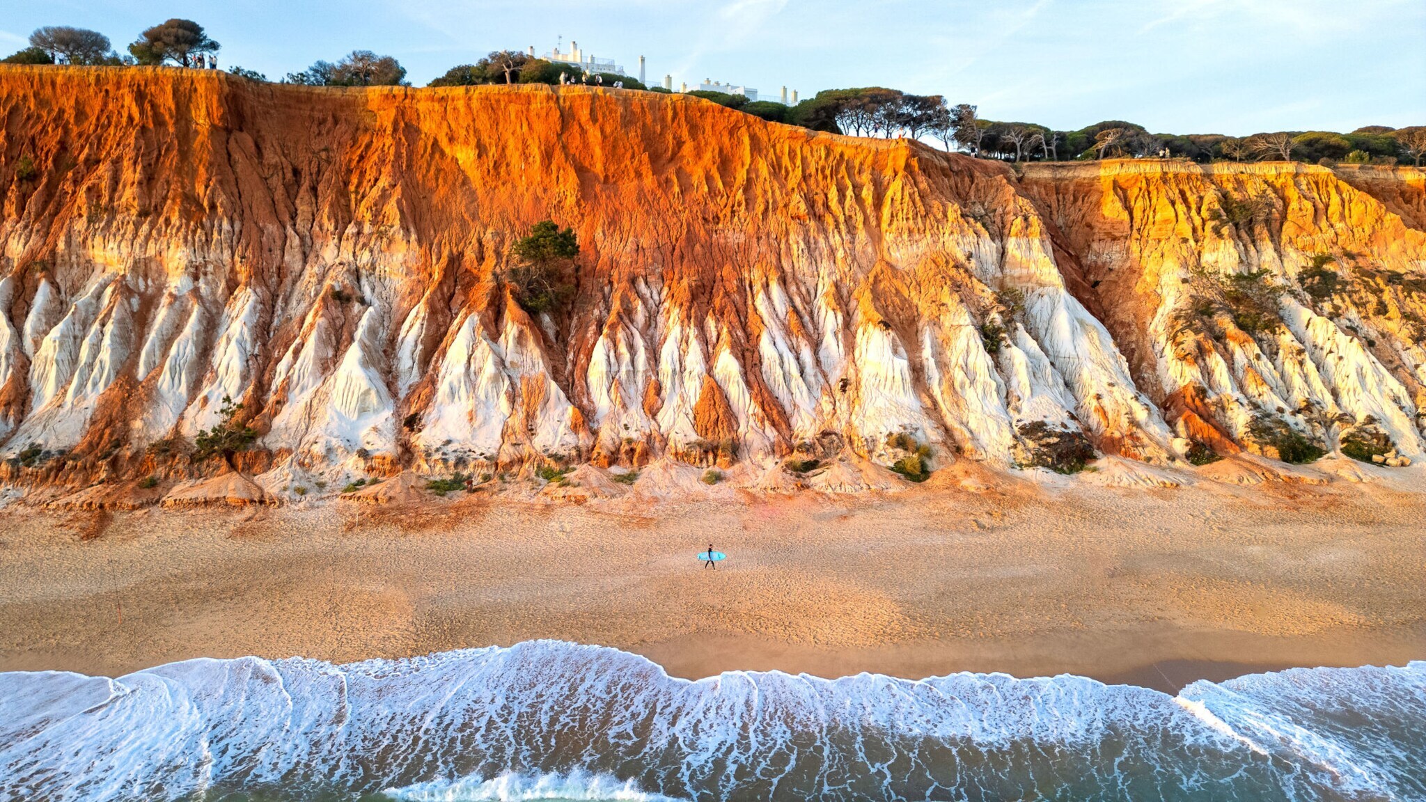 Hohe Klippen an der Praia da Falésia in Portugal, davor das Meer und der Strand, an dem eine Person mit Surfbrett entlangläuft.