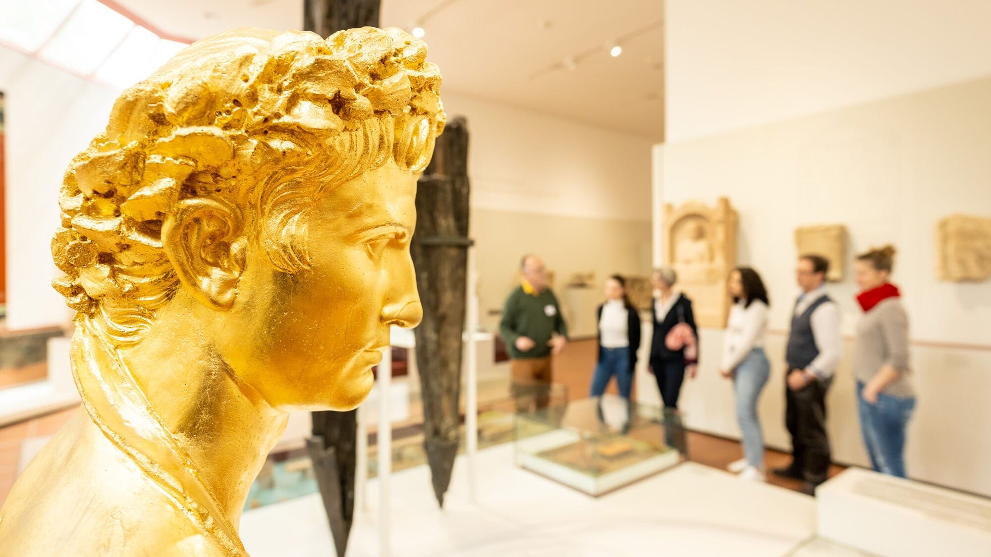 Goldene Büste mit lockigem Haar im Rheinischen Landesmuseum, im Hintergrund Besuchergruppe vor Ausstellungsstücken. Goldene Büste mit lockigem Haar im Rheinischen Landesmuseum, im Hintergrund Besuchergruppe vor Ausstellungsstücken.