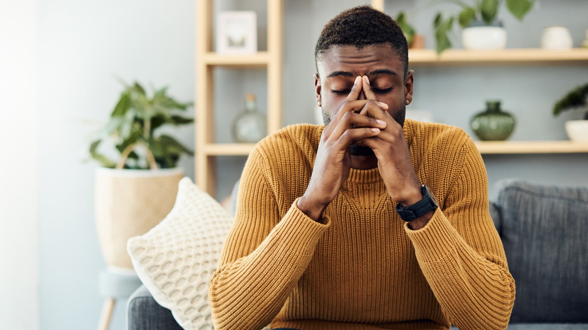 Ein Mann mit kurzem, dunklem Haar sitzt auf einem grauen Sofa, trägt einen orangefarbenen Pullover und schaut auf einen Laptop. Pflanzen und Regale im Hintergrund. Ein Mann mit kurzem, dunklem Haar sitzt auf einem grauen Sofa, trägt einen orangefarbenen Pullover und schaut auf einen Laptop. Pflanzen und Regale im Hintergrund.