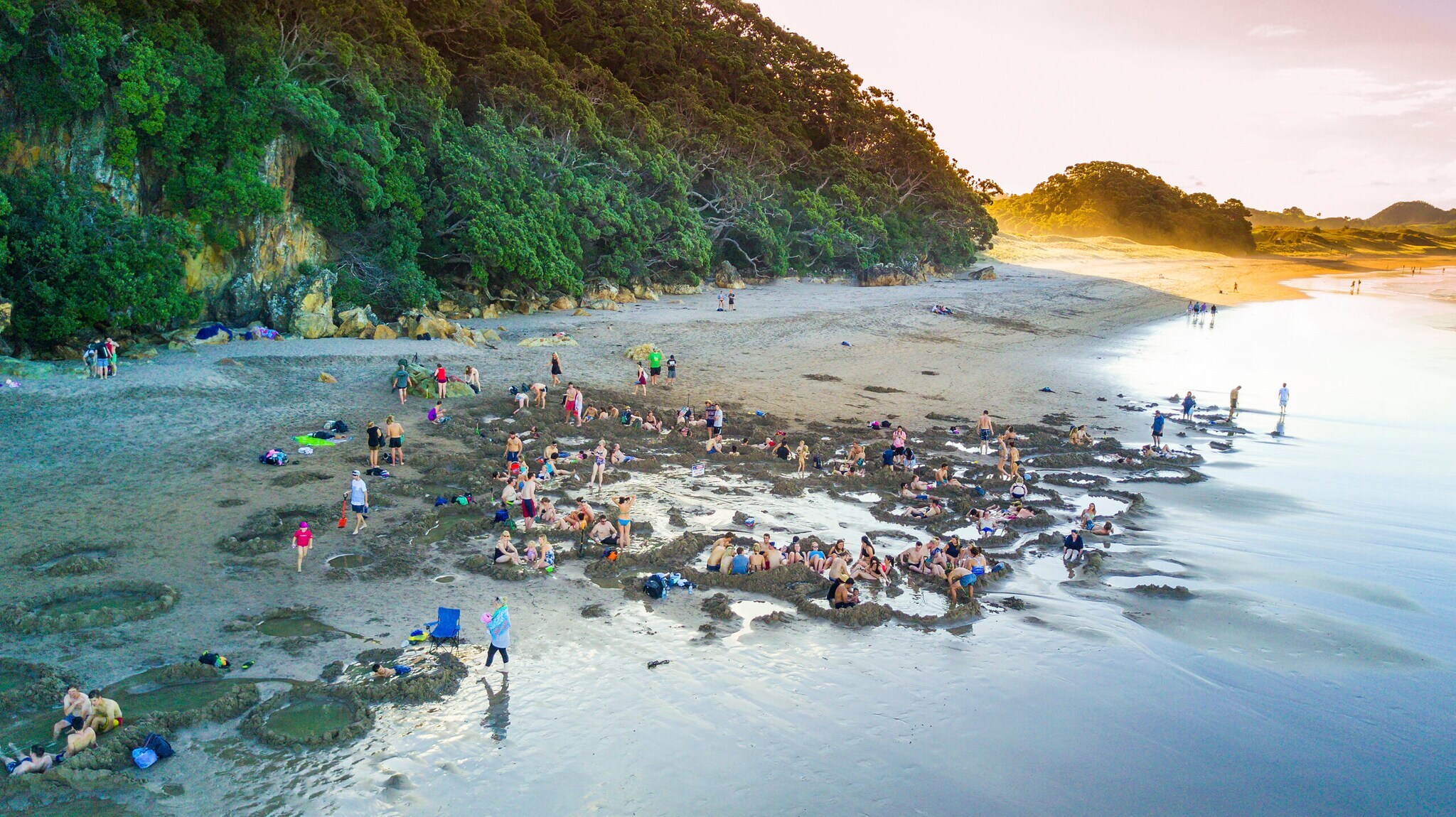 Ein Strand mit vielen sitzenden Menschen, im Hintergrund ein bewaldeter Felsen Ein Strand mit vielen sitzenden Menschen, im Hintergrund ein bewaldeter Felsen