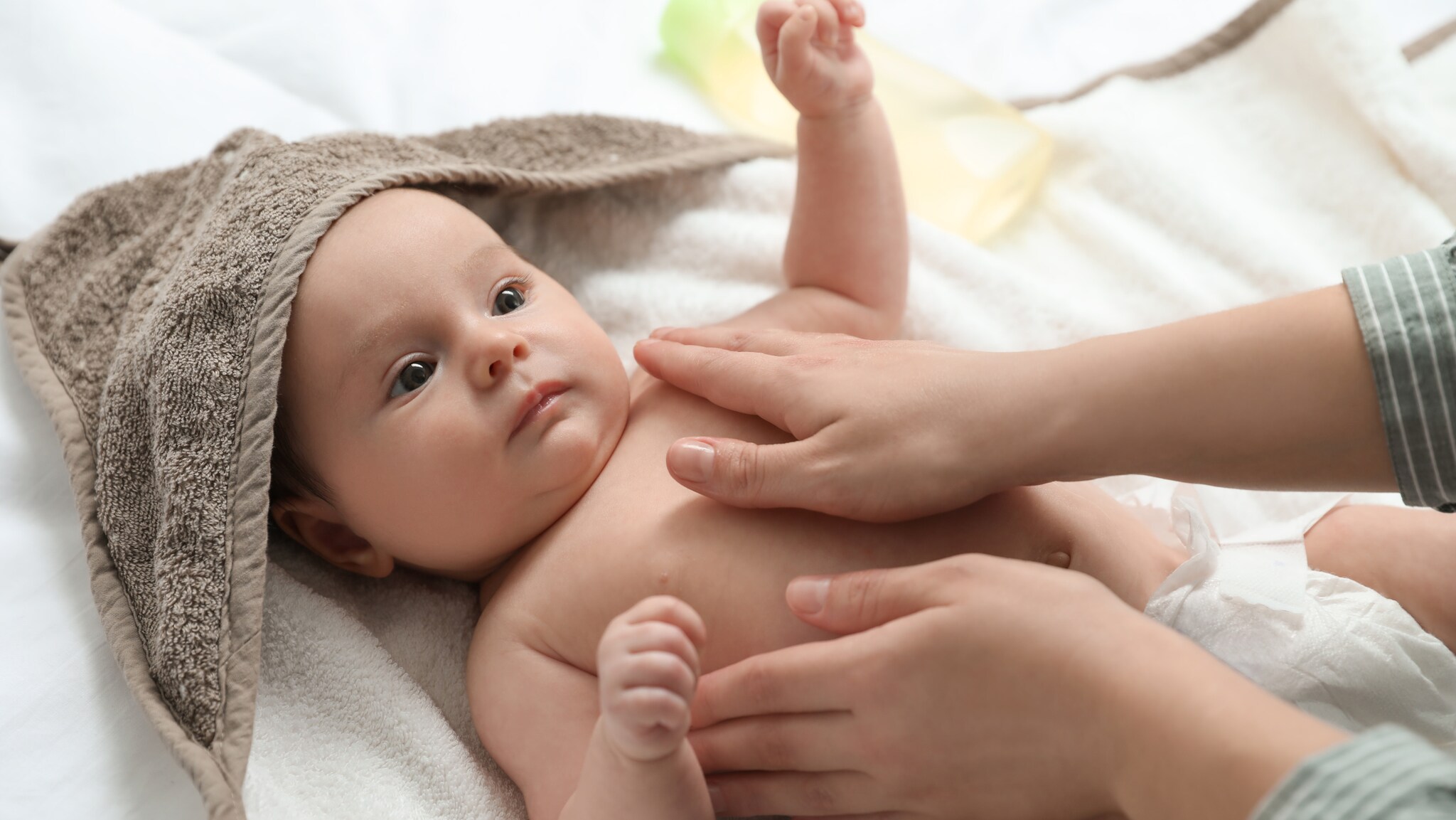 Baby liegt auf einer weißen Decke, teilweise mit braunem Handtuch bedeckt, während zwei Hände die Brust einreiben.