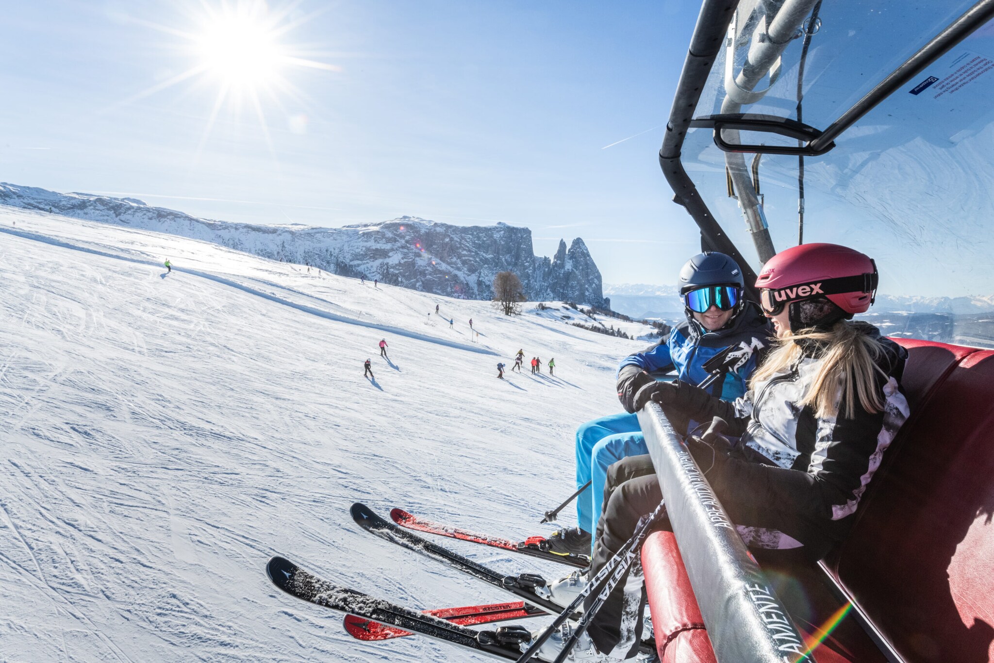 Ein Skifahrer und eine Skifahrerin sitzen in einem Sessellift über einer weitläufigen Piste vor Bergpanorama bei Sonnenschein