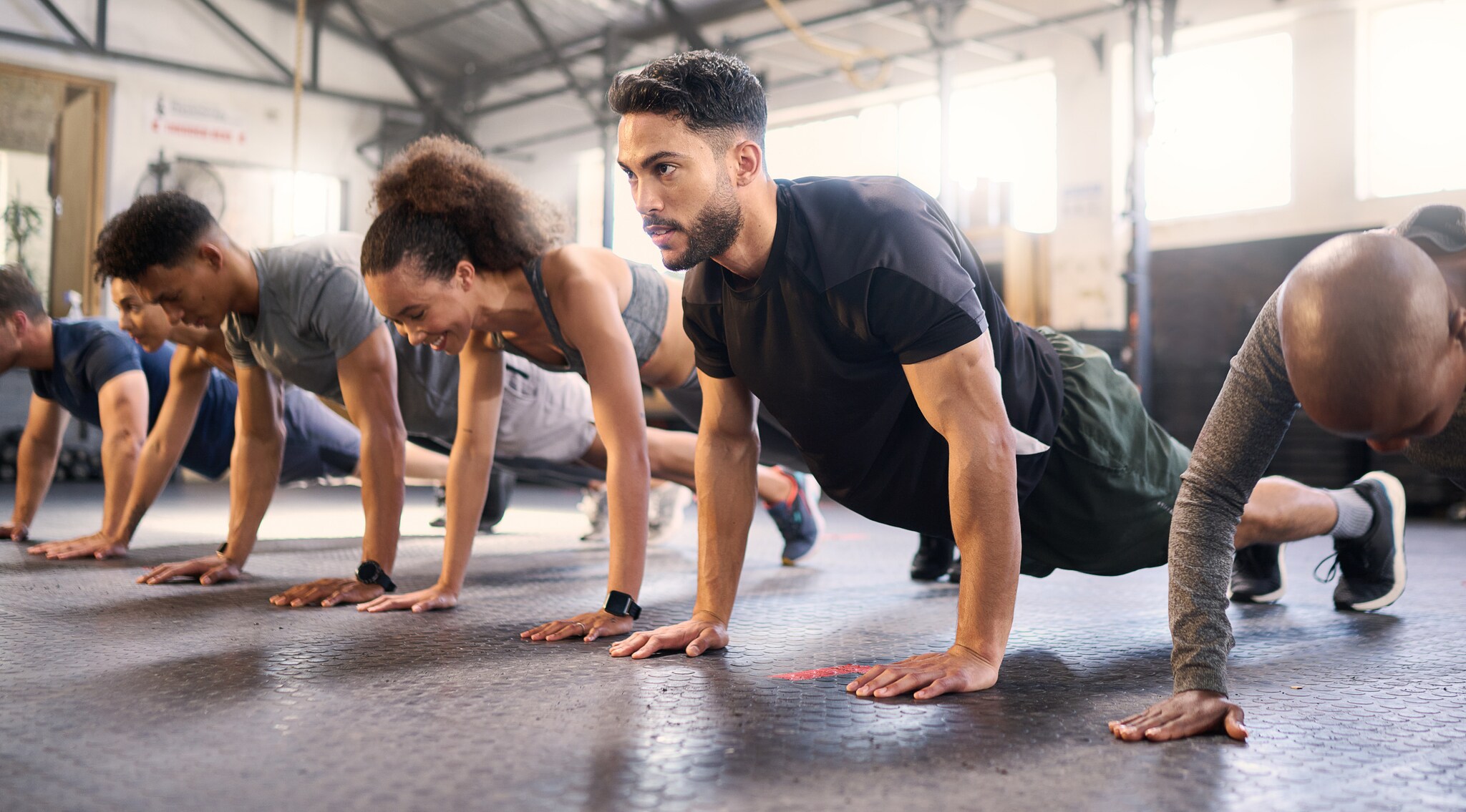 Mehrere Personen in Sportkleidung machen Liegestütze in einem hellen Fitnessstudio mit Gummiboden.