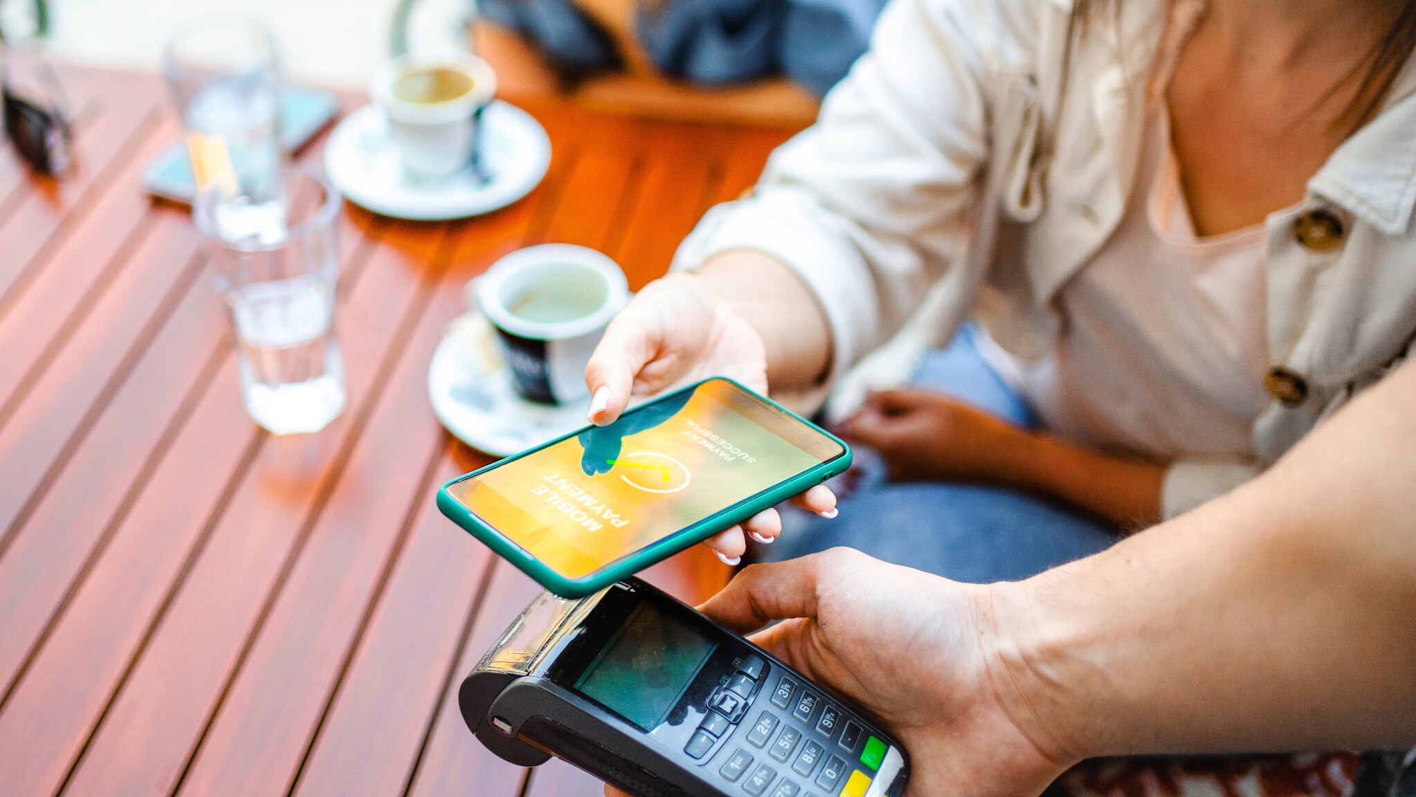 Nahaufnahme einer kontaktlosen Zahlung mit Kreditkarte via Smartphone an einem Kartenlesegerät an einem Cafétisch im Freien.