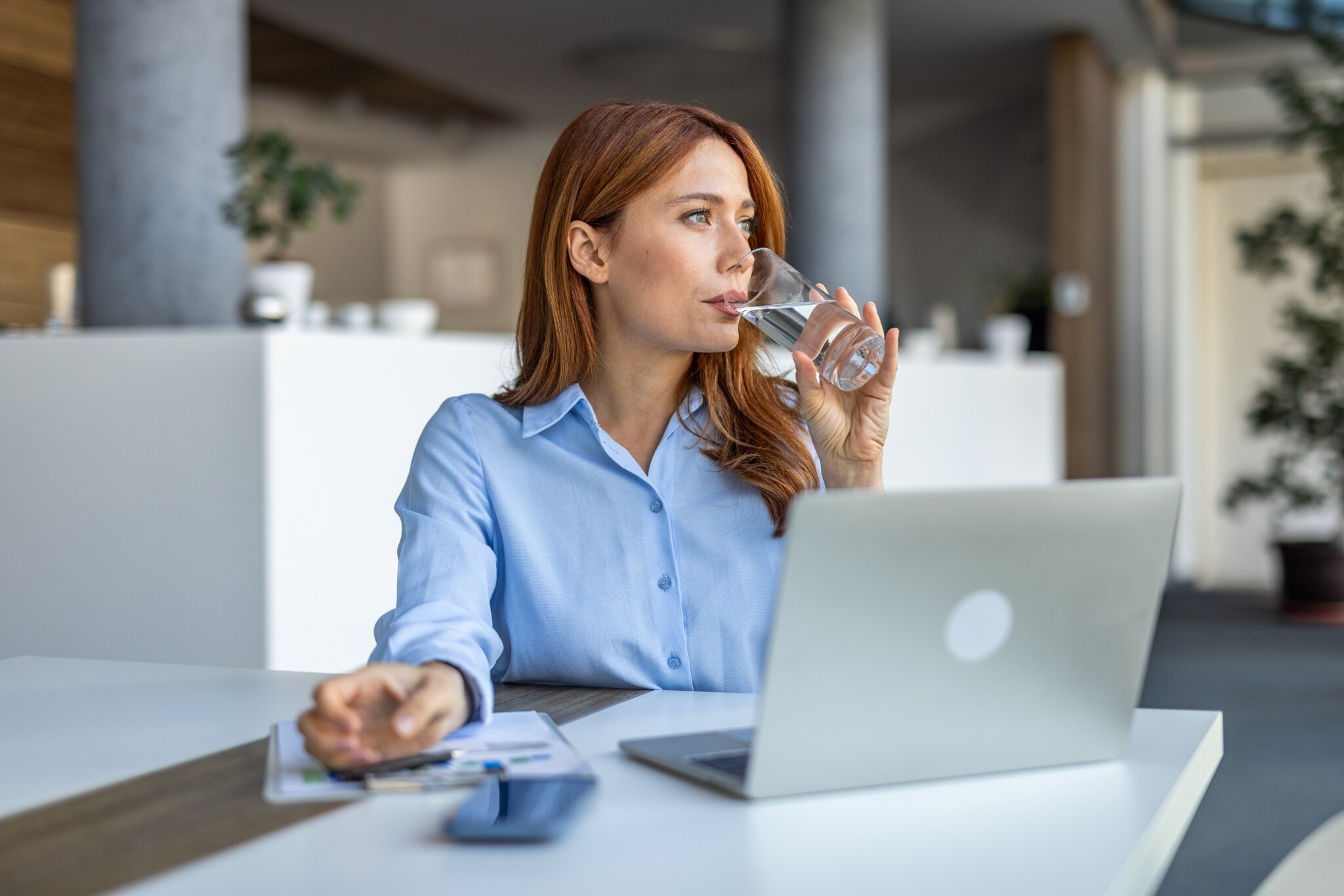 Eine Frau in hellblauer Bluse sitzt vor einem Laptop an einem Tisch und trinkt Wasser aus einem Glas.