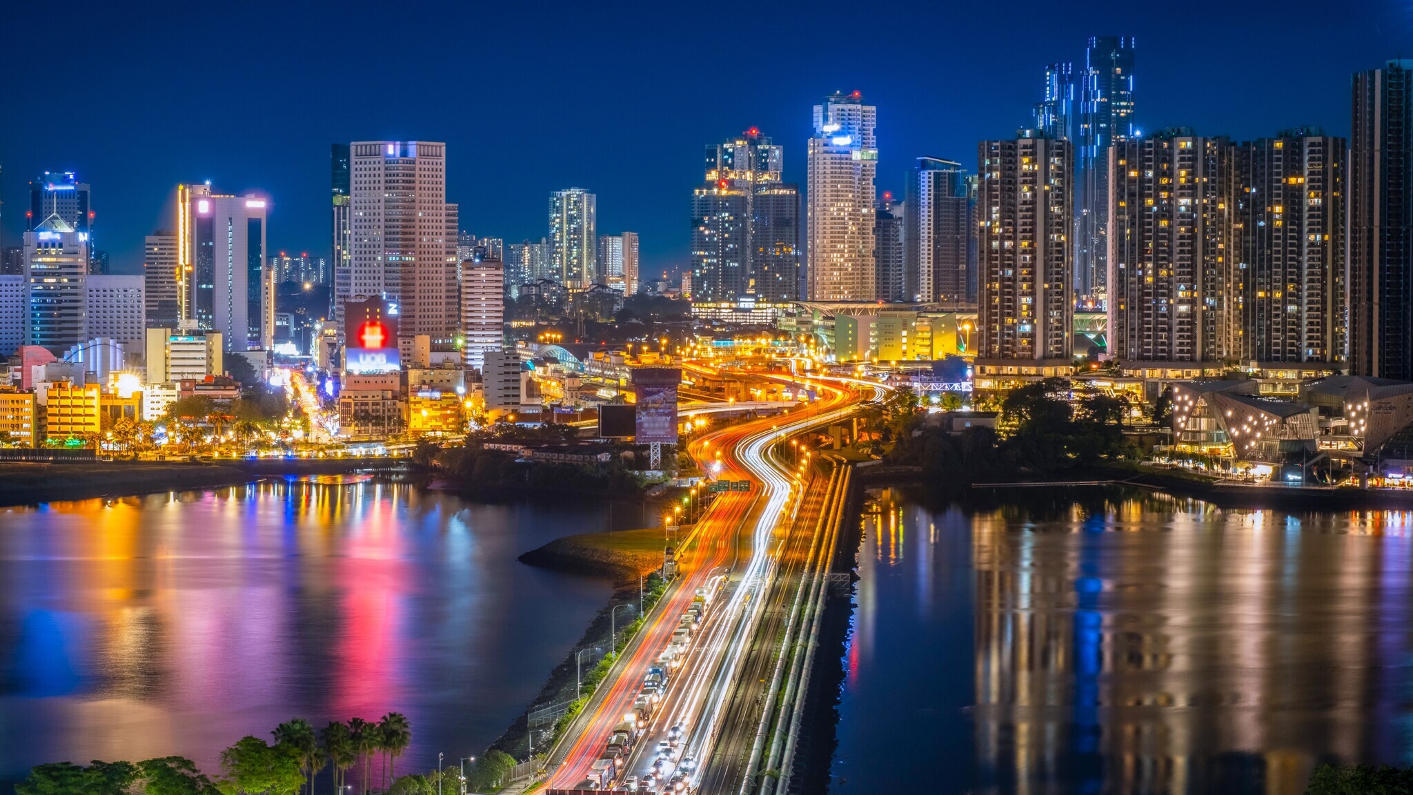 Nächtliche Stadtansicht mit beleuchteten Hochhäusern und einer vielbefahrenen Brücke über Wasser, Lichtspuren von Fahrzeugen sichtbar. Nächtliche Stadtansicht mit beleuchteten Hochhäusern und einer vielbefahrenen Brücke über Wasser, Lichtspuren von Fahrzeugen sichtbar.