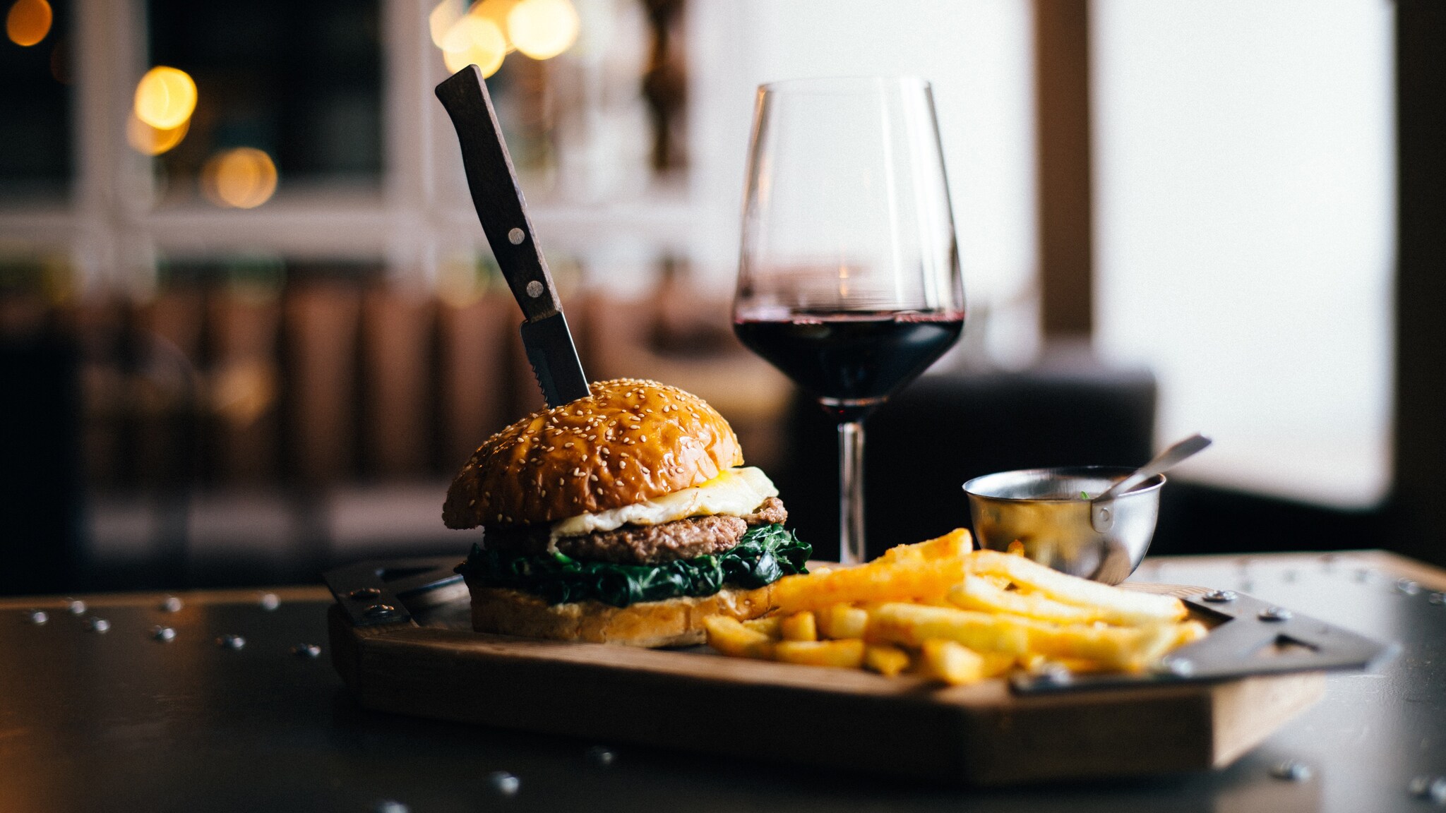 Burger mit Sesambrötchen, Spiegelei und Spinat, Messer steckt im Burger, daneben Pommes und ein Glas Rotwein auf einem Holzbrett