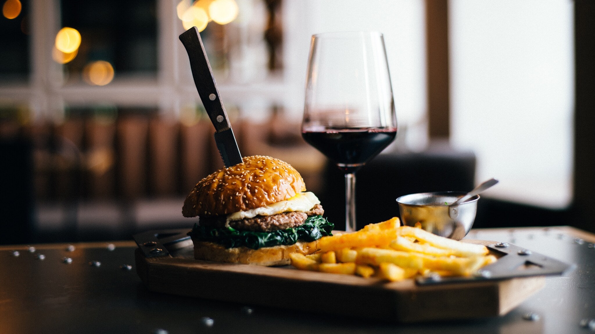 Burger mit Sesambrötchen, Spiegelei und Spinat, Messer steckt im Burger, daneben Pommes und ein Glas Rotwein auf einem Holzbrett Burger mit Sesambrötchen, Spiegelei und Spinat, Messer steckt im Burger, daneben Pommes und ein Glas Rotwein auf einem Holzbrett