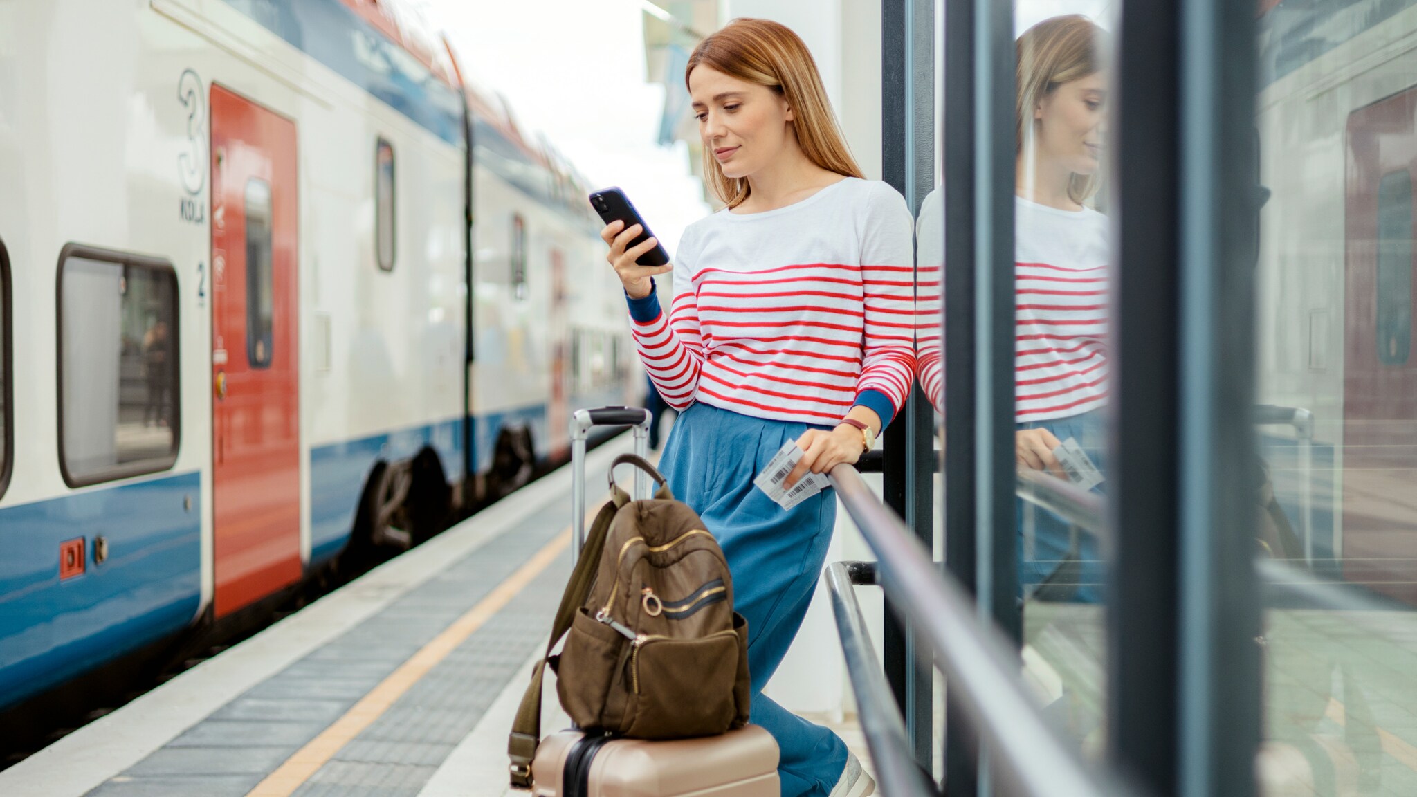 Eine Frau in weißem Shirt mit roten Streifen und blauer Hose lehnt an einer Glaswand am Bahnsteig mit Smartphone in der Hand, neben ihr ein Koffer.