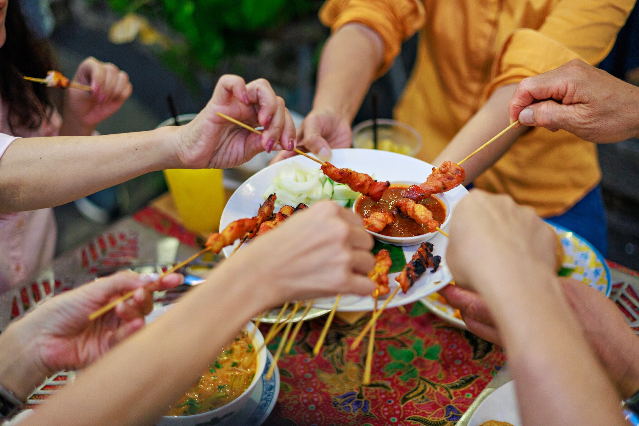 Mehrere Hände dippen Hühnchenspieße in eine braune Soße auf einem Teller in ihrer Mitte an einem Tisch.