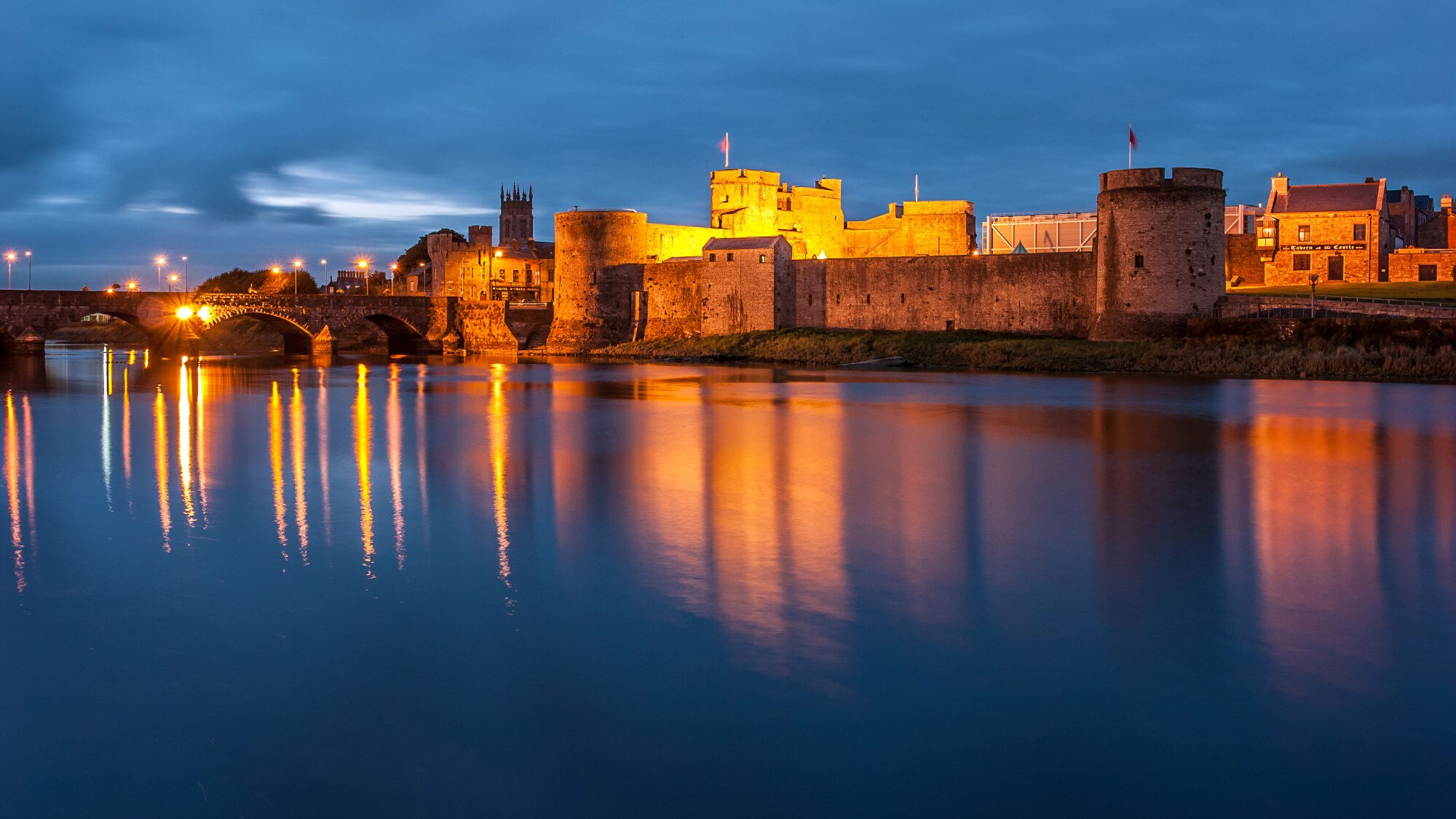 Blick auf eine beleuchtete Burg am Wasser bei Dämmerung. Die Burg hat Türme und eine Brücke im Vordergrund. Reflektionen im Wasser sind sichtbar. Blick auf eine beleuchtete Burg am Wasser bei Dämmerung. Die Burg hat Türme und eine Brücke im Vordergrund. Reflektionen im Wasser sind sichtbar.
