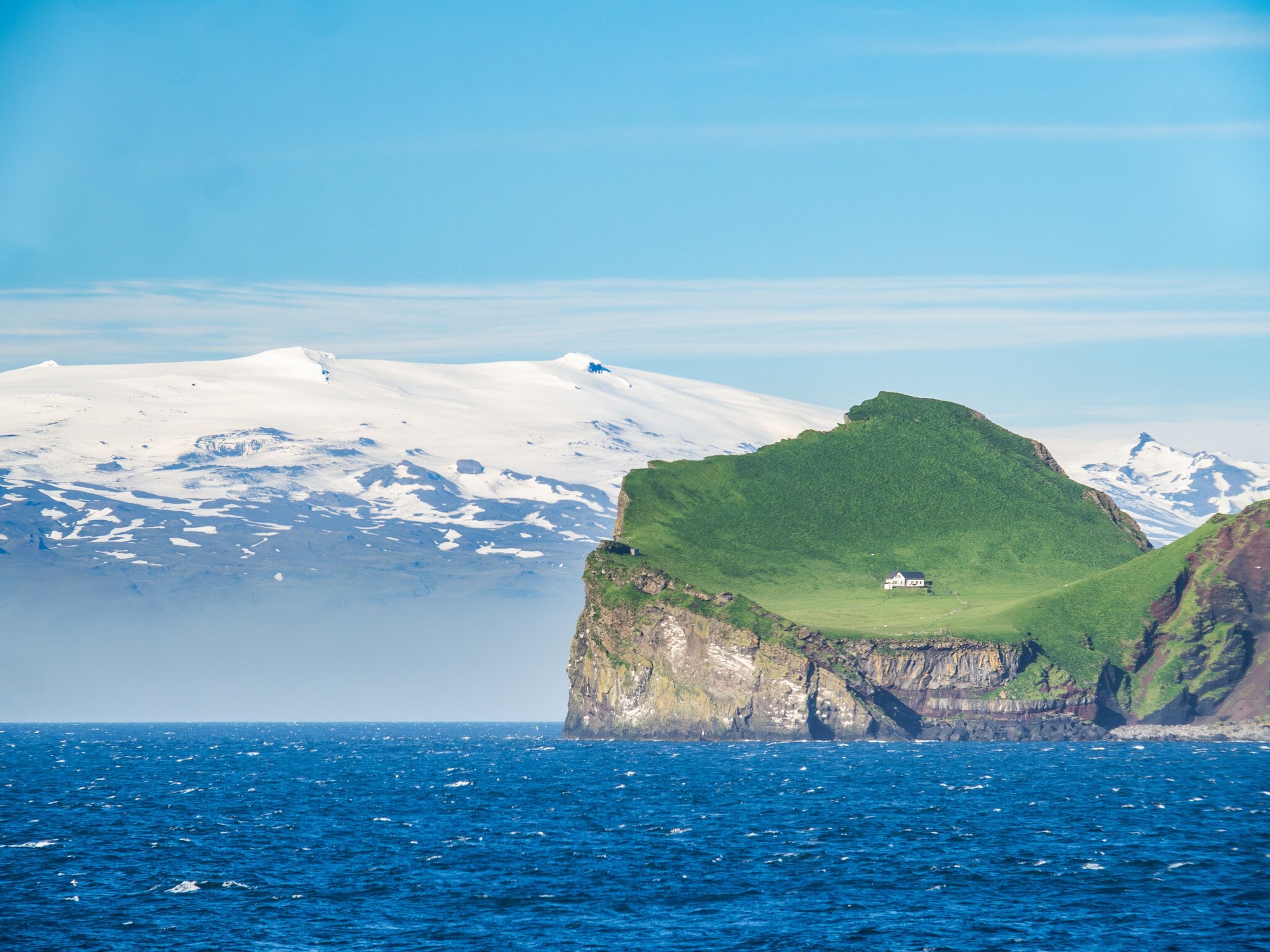 Blick auf die grüne isländische Insel Ellidaey mit einer einsamen Jagdhütte darauf und dem, unter einem Gletscher liegenden Vulkan Eyjafjallajökull im Hintergrund. Blick auf die grüne isländische Insel Ellidaey mit einer einsamen Jagdhütte darauf und dem, unter einem Gletscher liegenden Vulkan Eyjafjallajökull im Hintergrund.