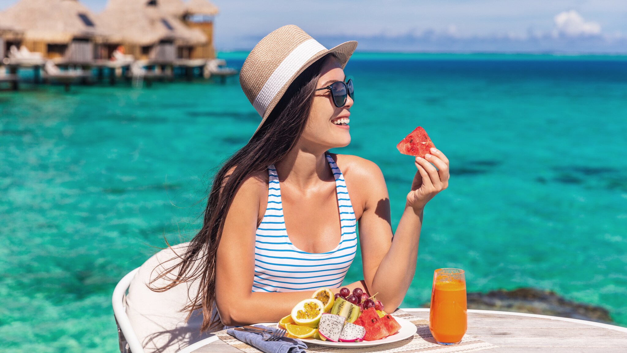 Eine Frau mit Sonnenhut sitzt an einem Tisch mit Obstteller und Fruchtsaft vor türkisblauem Meer mit Überwasser-Bungalows.