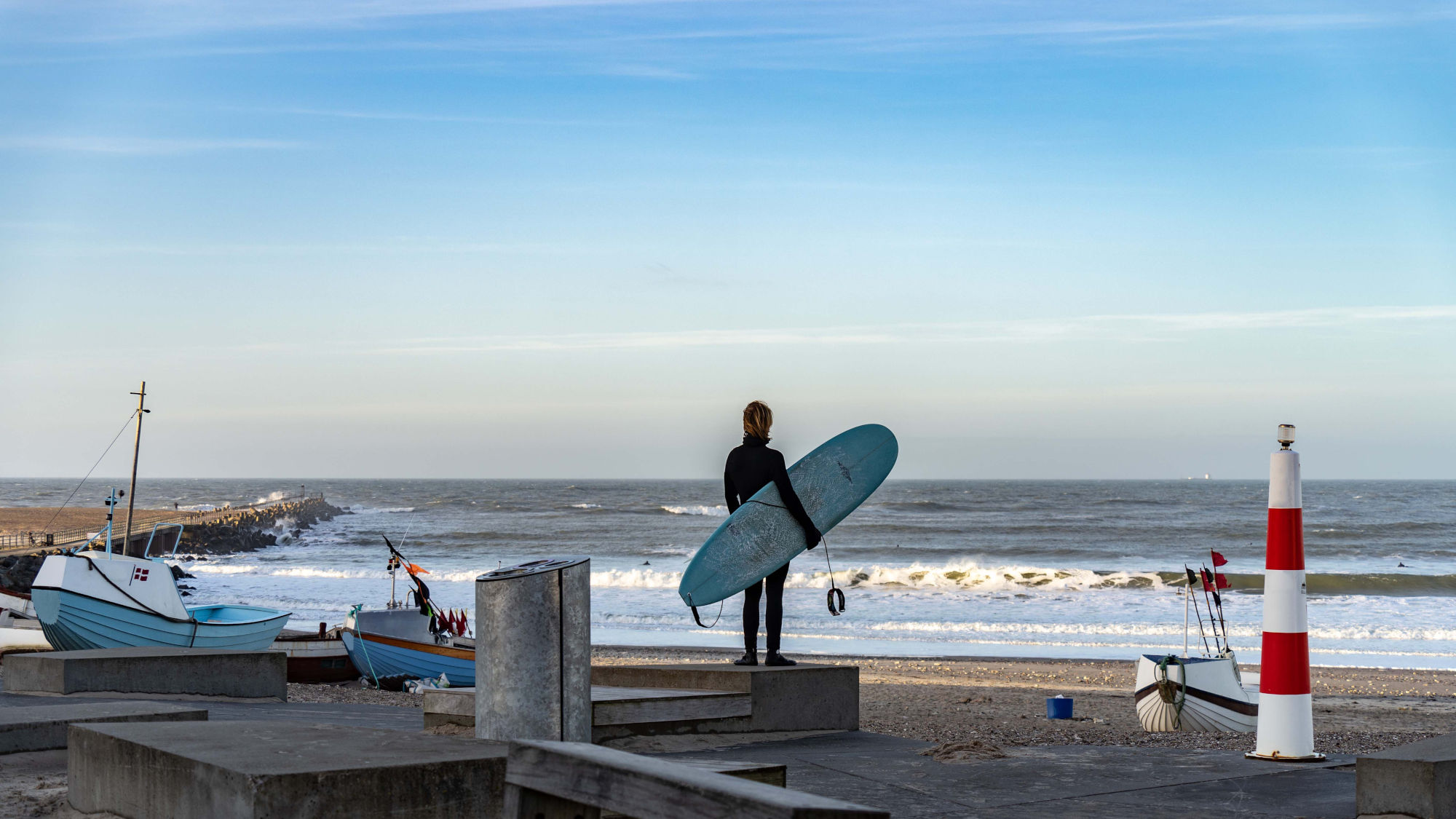 Surfer mit blauem Surfbrett steht am Strand von Dänemark und blickt auf die Nordsee, im Hintergrund Boote und ein rot-weiß gestreifter Leuchtturm.