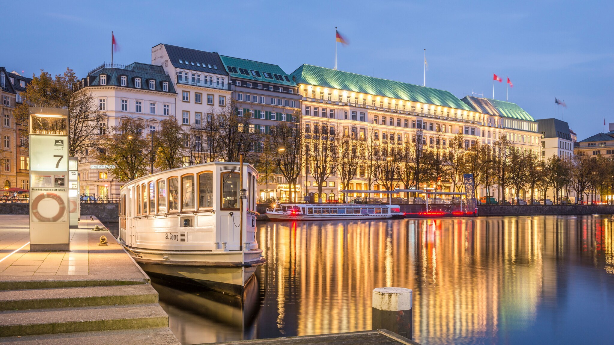Beleuchtete Häuserfront im hanseatischen Stil mit Luxushotels an einer Promenade mit Spieglungen im Wasser bei Abenddämmerung, im Vordergrund eine Barkasse an einem Bootsanleger.