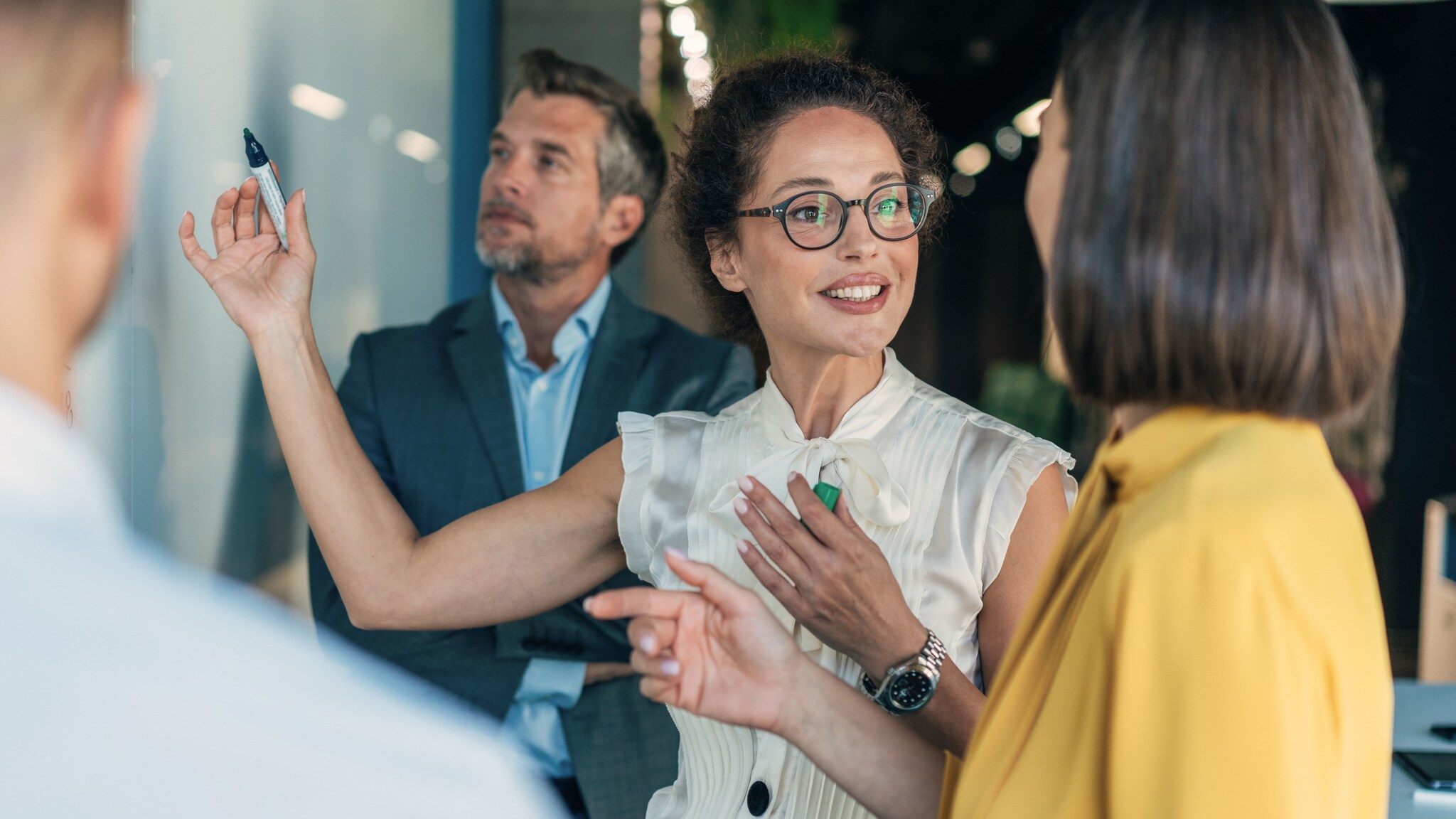 Vier Personen in Business-Kleidung stehen vor einer Wand und kommunizieren, eine Frau hält einen Stift in der Hand hoch.