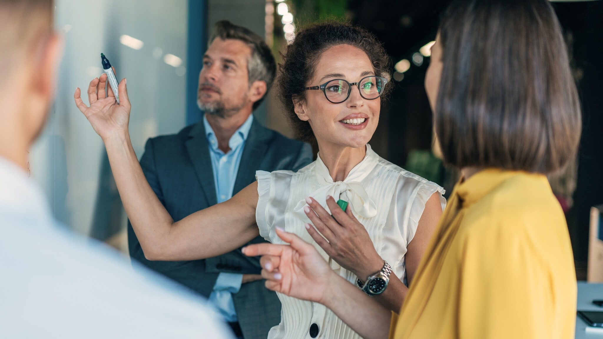 Vier Personen in Business-Kleidung stehen vor einer Wand und kommunizieren, eine Frau hält einen Stift in der Hand hoch. Vier Personen in Business-Kleidung stehen vor einer Wand und kommunizieren, eine Frau hält einen Stift in der Hand hoch.