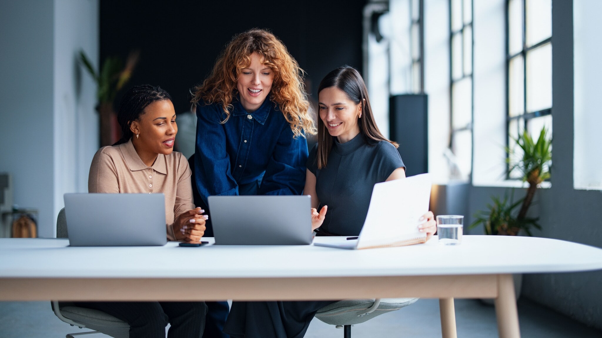 Drei Frauen schauen gemeinsam auf ein Laptop auf einem großen Tisch mit zwei weiteren Laptops in einem modernen Büro. Drei Frauen schauen gemeinsam auf ein Laptop auf einem großen Tisch mit zwei weiteren Laptops in einem modernen Büro.