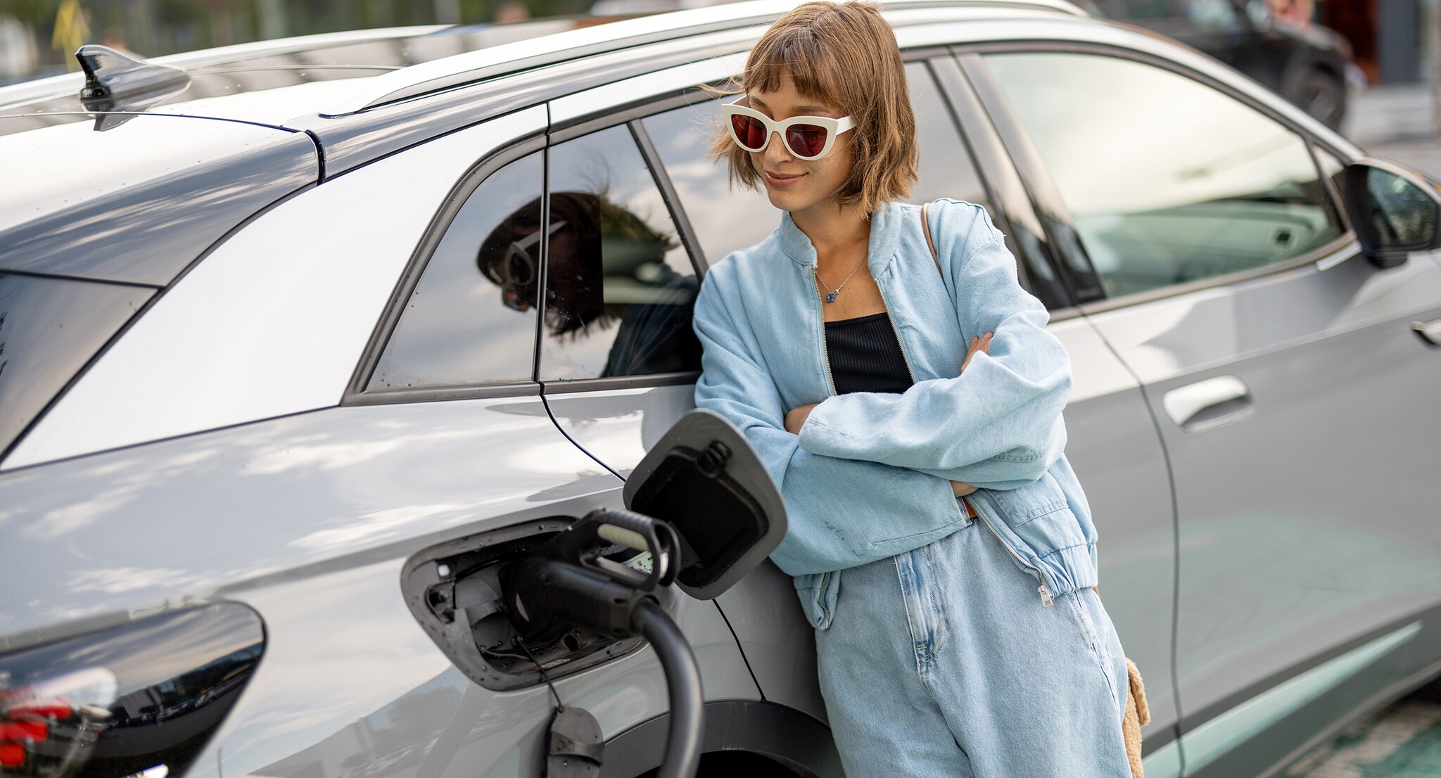 Portrait of a young woman standing near her electric car charging on a public station. Concept of modern lifestyle and success