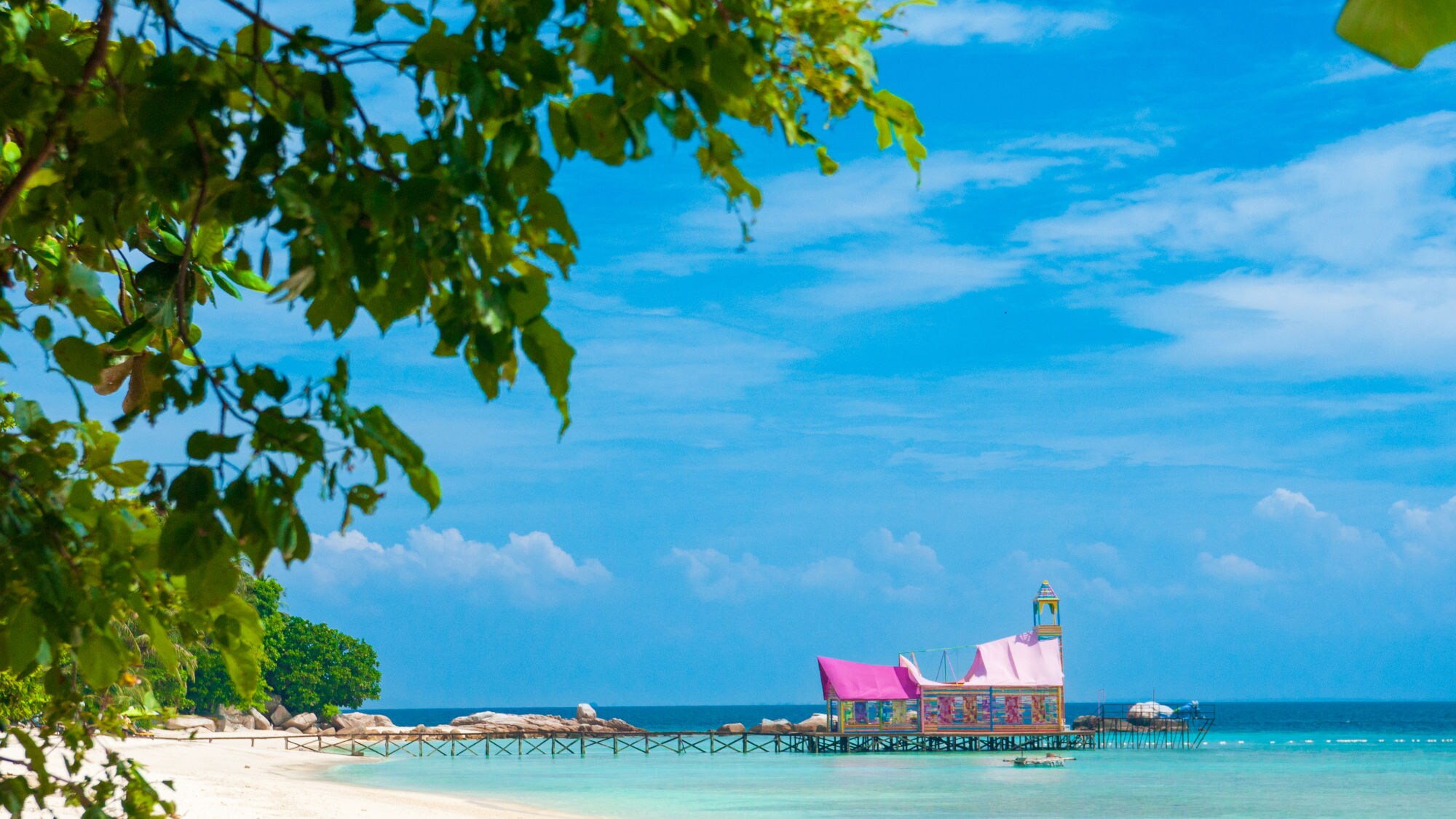 Strand mit grünem Laub im Vordergrund, hölzerner Steg mit buntem Pavillon über türkisfarbenem Meer bei blauem Himmel. Strand mit grünem Laub im Vordergrund, hölzerner Steg mit buntem Pavillon über türkisfarbenem Meer bei blauem Himmel.