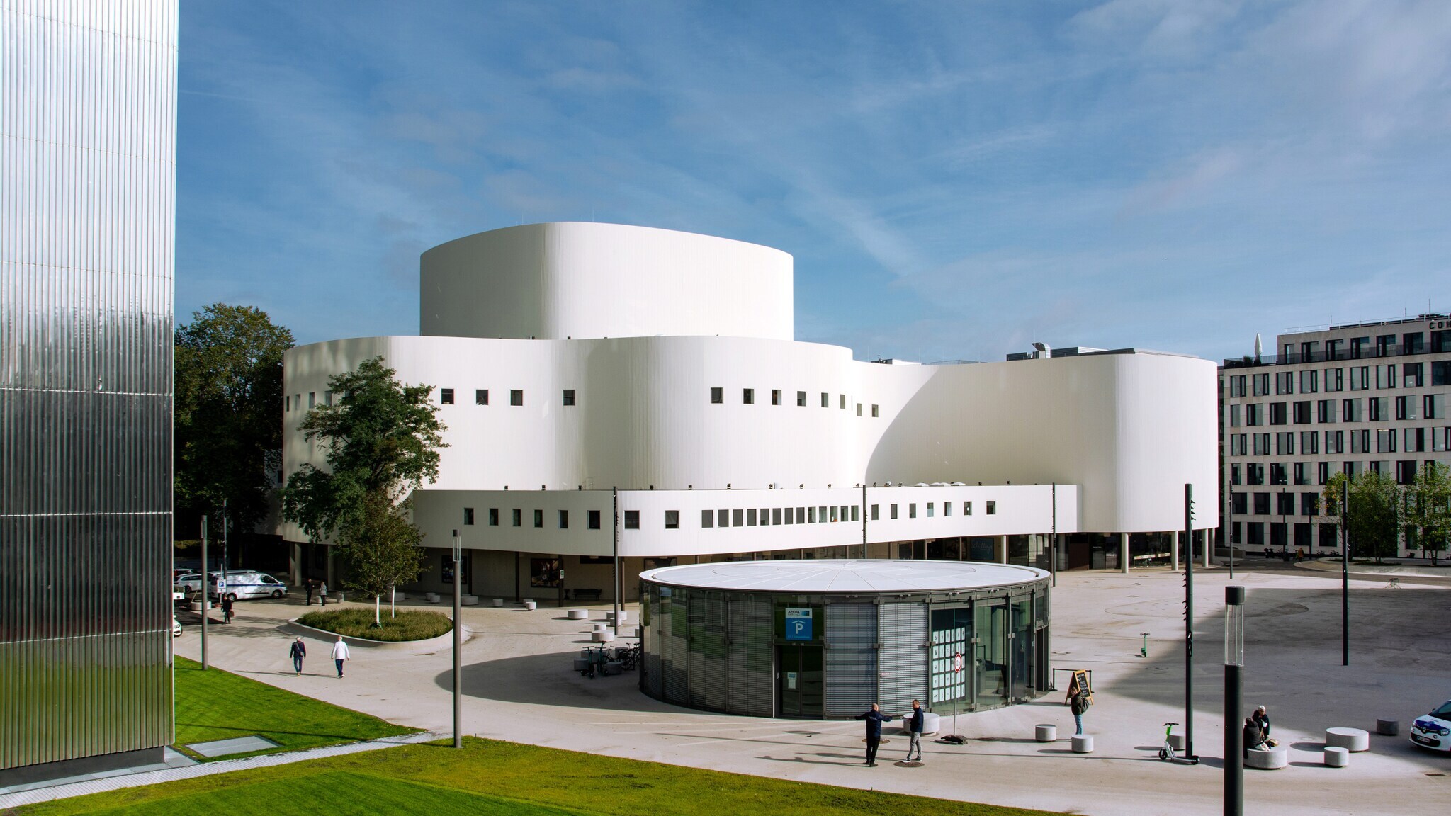 Modernes weißes Theatergebäude mit runden Formen und kleinen quadratischen Fenstern, umgeben von einem Platz mit wenigen Menschen und Autos Modernes weißes Theatergebäude mit runden Formen und kleinen quadratischen Fenstern, umgeben von einem Platz mit wenigen Menschen und Autos