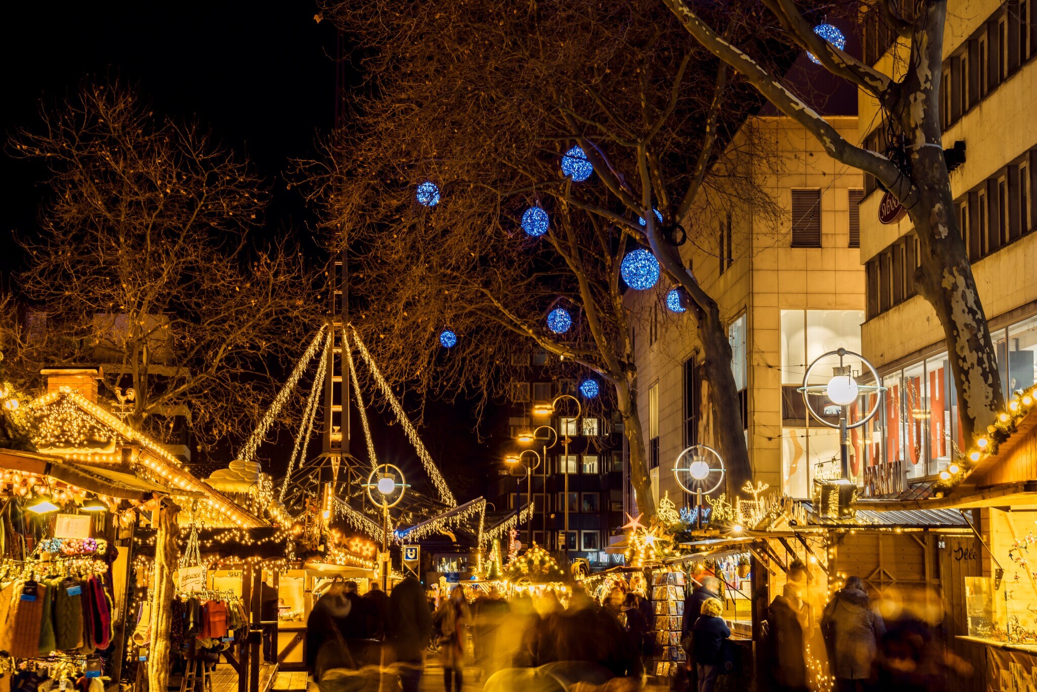 Beleuchtete Buden in einer Einkaufsmeile, an den Bäumen hängen blaue Lichtkugeln