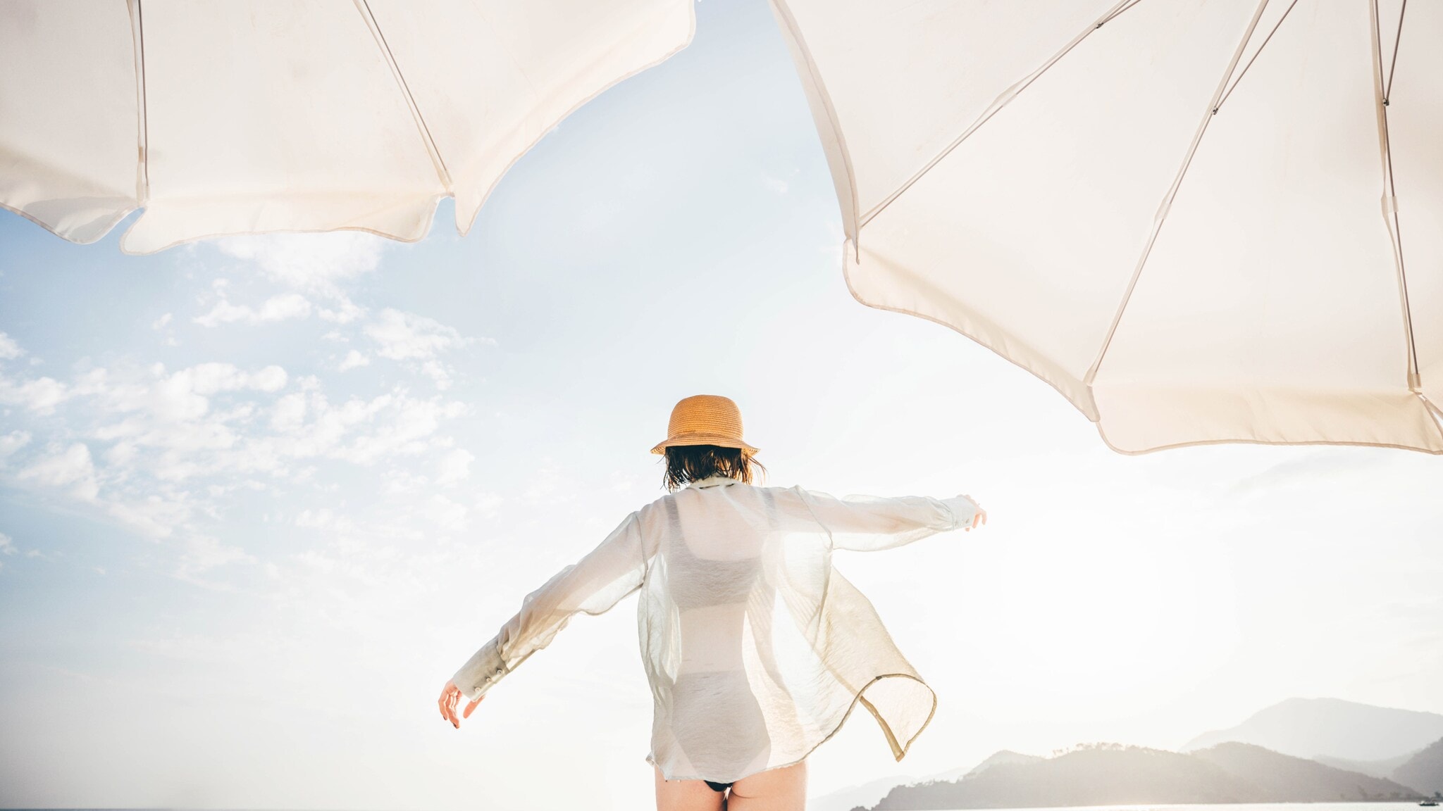 Eine Person mit einem Hut und einem weißen Oberteil steht mit dem Rücken zum Betrachter unter zwei Sonnenschirmen am Strand. Der Himmel ist teilweise bewölkt. Eine Person mit einem Hut und einem weißen Oberteil steht mit dem Rücken zum Betrachter unter zwei Sonnenschirmen am Strand. Der Himmel ist teilweise bewölkt.
