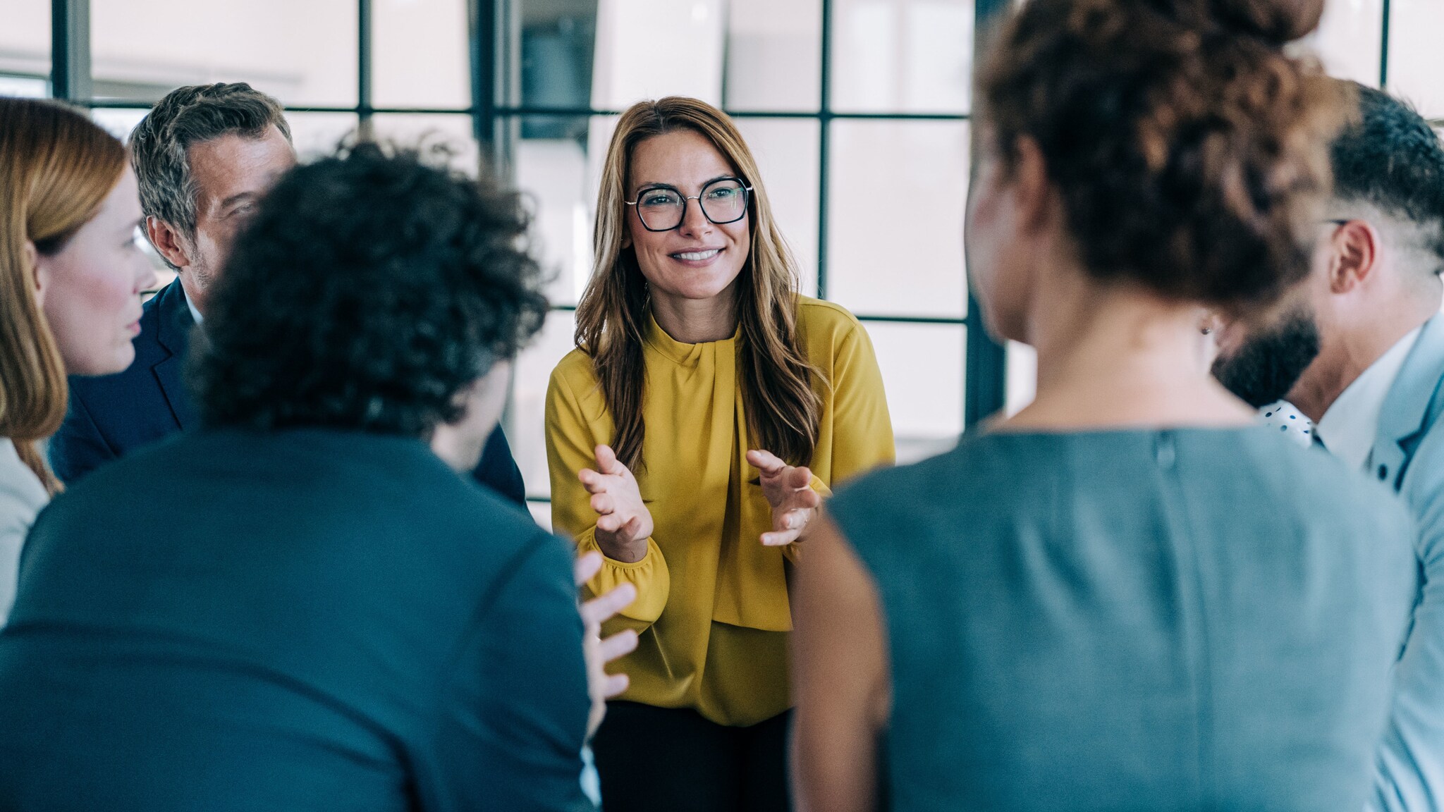 Mehrere Personen in Business-Kleidung stehen in einem Kreis, im Zentrum eine lächelnde, gestikulierende Frau im gelben Oberteil. Mehrere Personen in Business-Kleidung stehen in einem Kreis, im Zentrum eine lächelnde, gestikulierende Frau im gelben Oberteil.