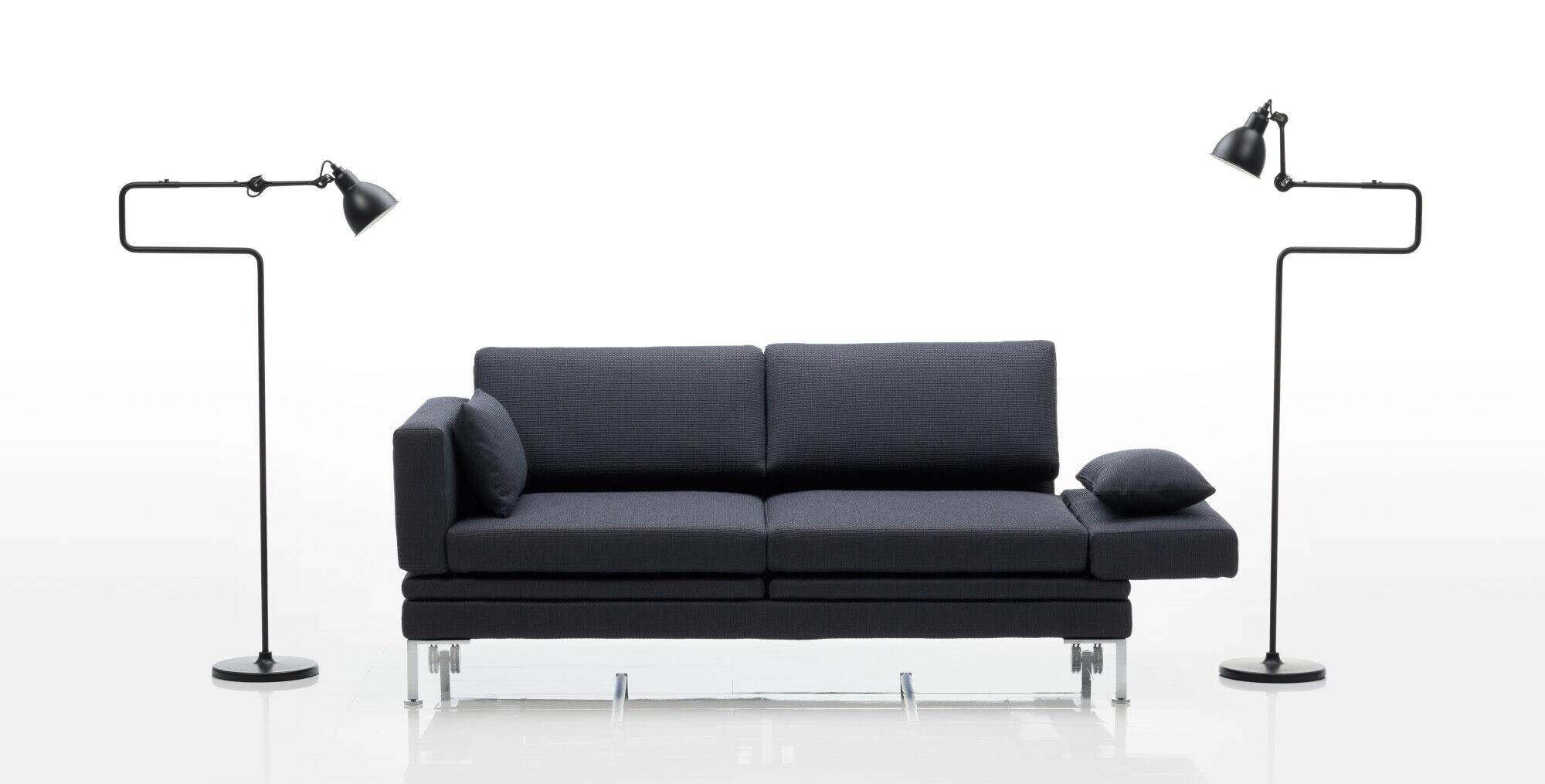 Ein geradliniges, ausklappbares Sofa mit anthrazitfarbenen Bezug, daneben zwei schwarze Stehleuchten im industriellen Design vor weißem Hintergrund
