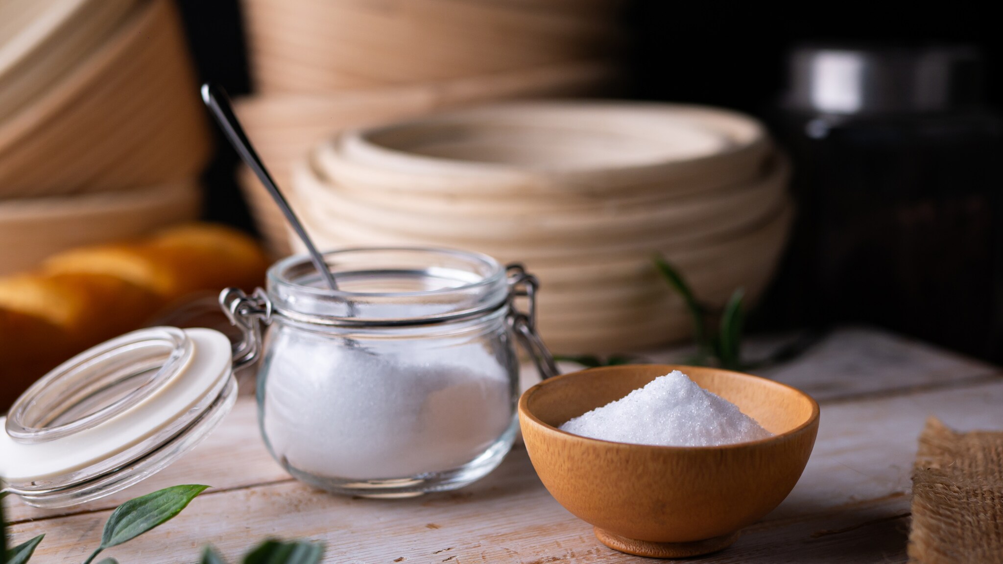 Auf einem Holztisch steht eine hellbraune Schale mit dem weißen Xylit, daneben steht ein Einmachglas mit Löffel und enthält ebenfalls Xylit.