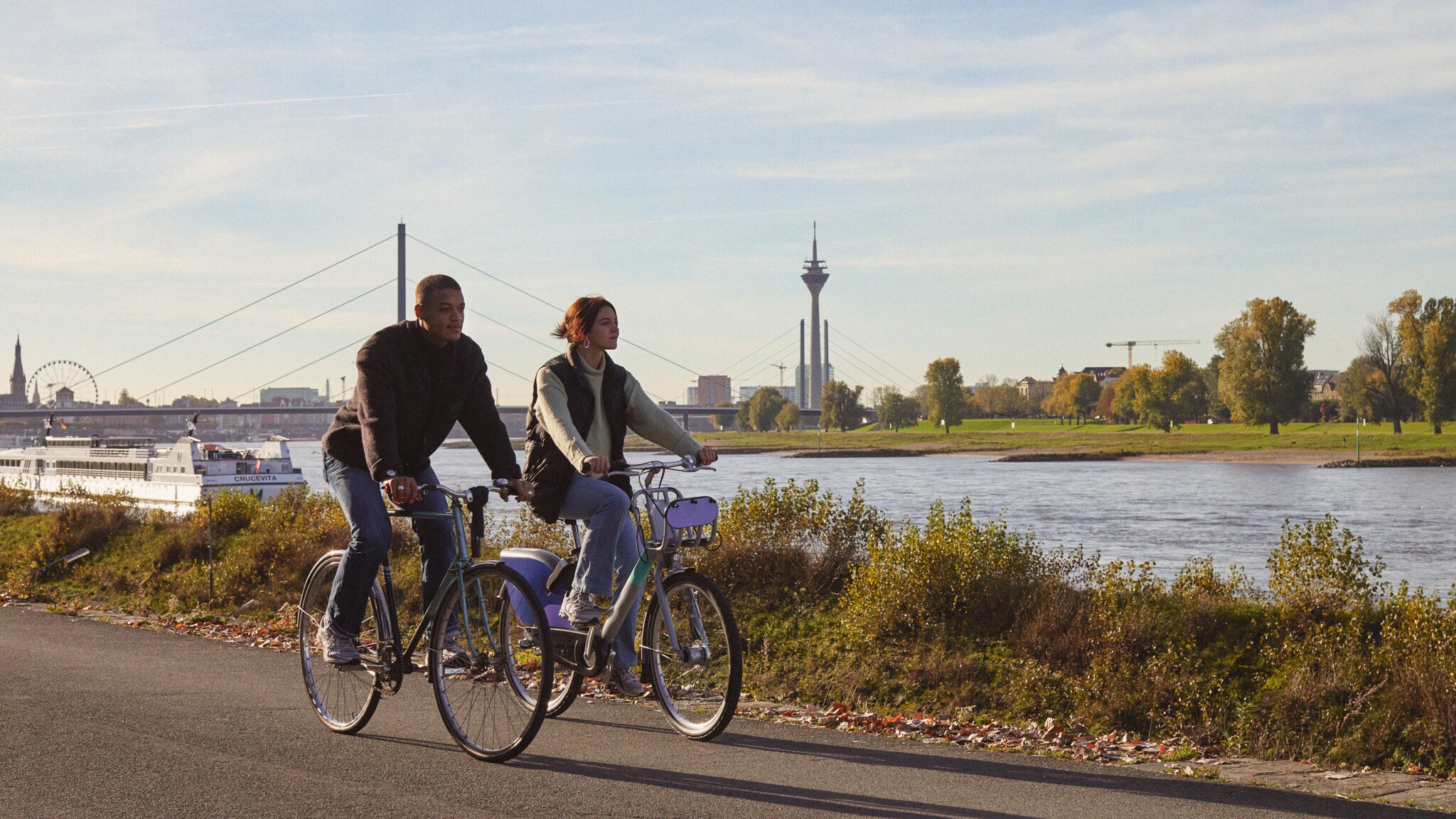 Zwei Personen auf Fahrrädern fahren am Ufer eines Flusses entlang. Im Hintergrund sind Bäume, ein Fernsehturm und ein Riesenrad sichtbar.