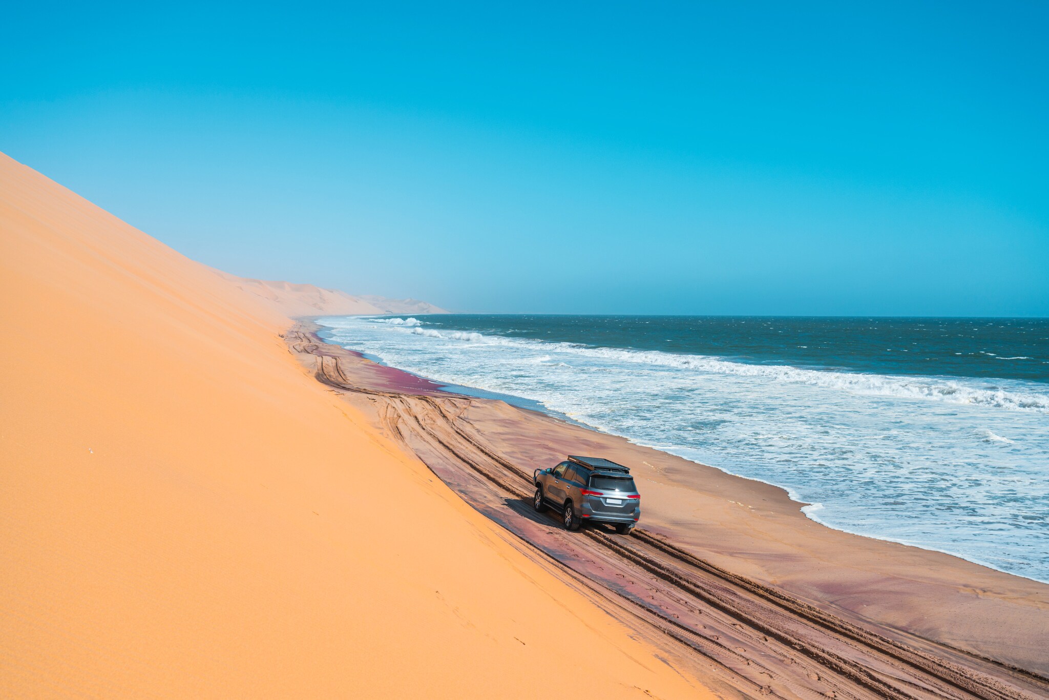 Ein Jeep fährt am Fuße einer Düne eine Meeresküste entlang. Ein Jeep fährt am Fuße einer Düne eine Meeresküste entlang.