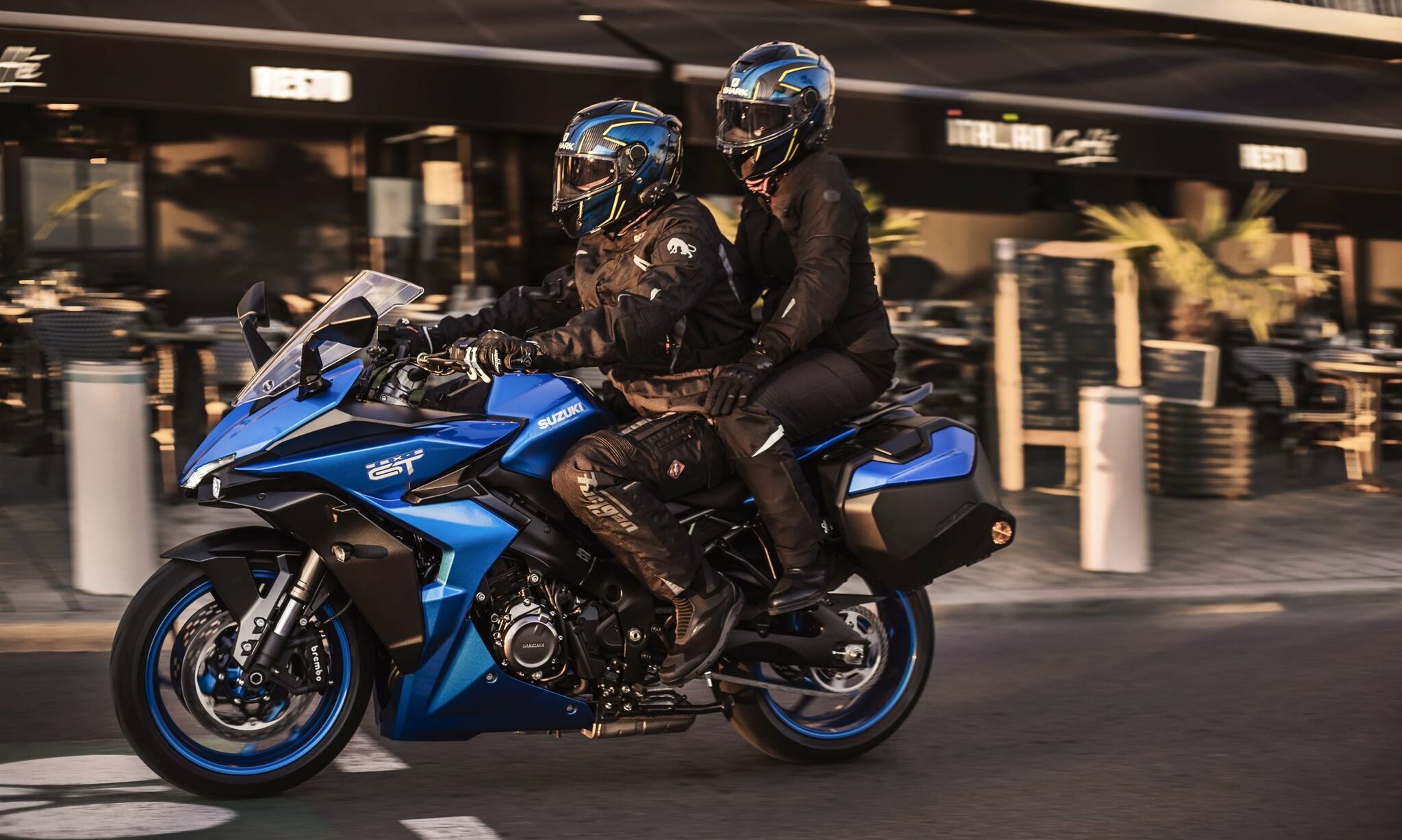 Eine blaue Suzuki GSX-S1000GT mit Fahrer und Sozia rollt vor einem Bistro vorbei Eine blaue Suzuki GSX-S1000GT mit Fahrer und Sozia rollt vor einem Bistro vorbei