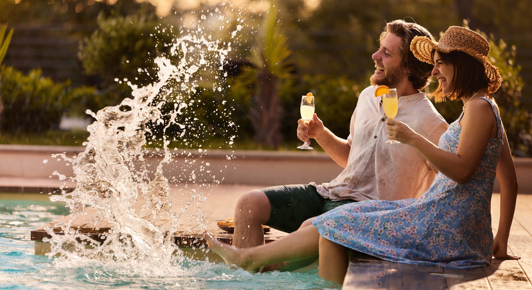Eine Frau und ein Mann sitzen am Rand eines Pools mit einem Getränk in der Hand und spritzen mit ihren Füßen.