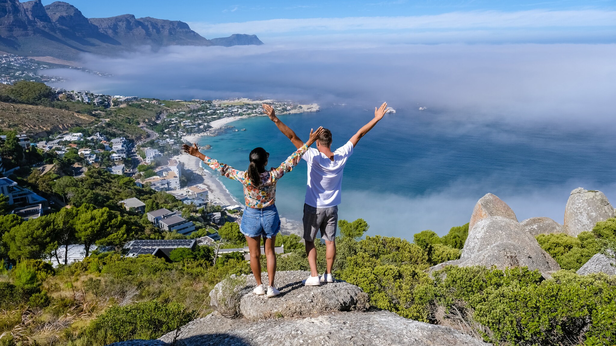 Zwei Personen stehen auf einem Felsen mit erhobenen Armen und blicken auf eine Küstenlandschaft mit Meer, Häusern und Bergen unter blauem Himmel. Zwei Personen stehen auf einem Felsen mit erhobenen Armen und blicken auf eine Küstenlandschaft mit Meer, Häusern und Bergen unter blauem Himmel.