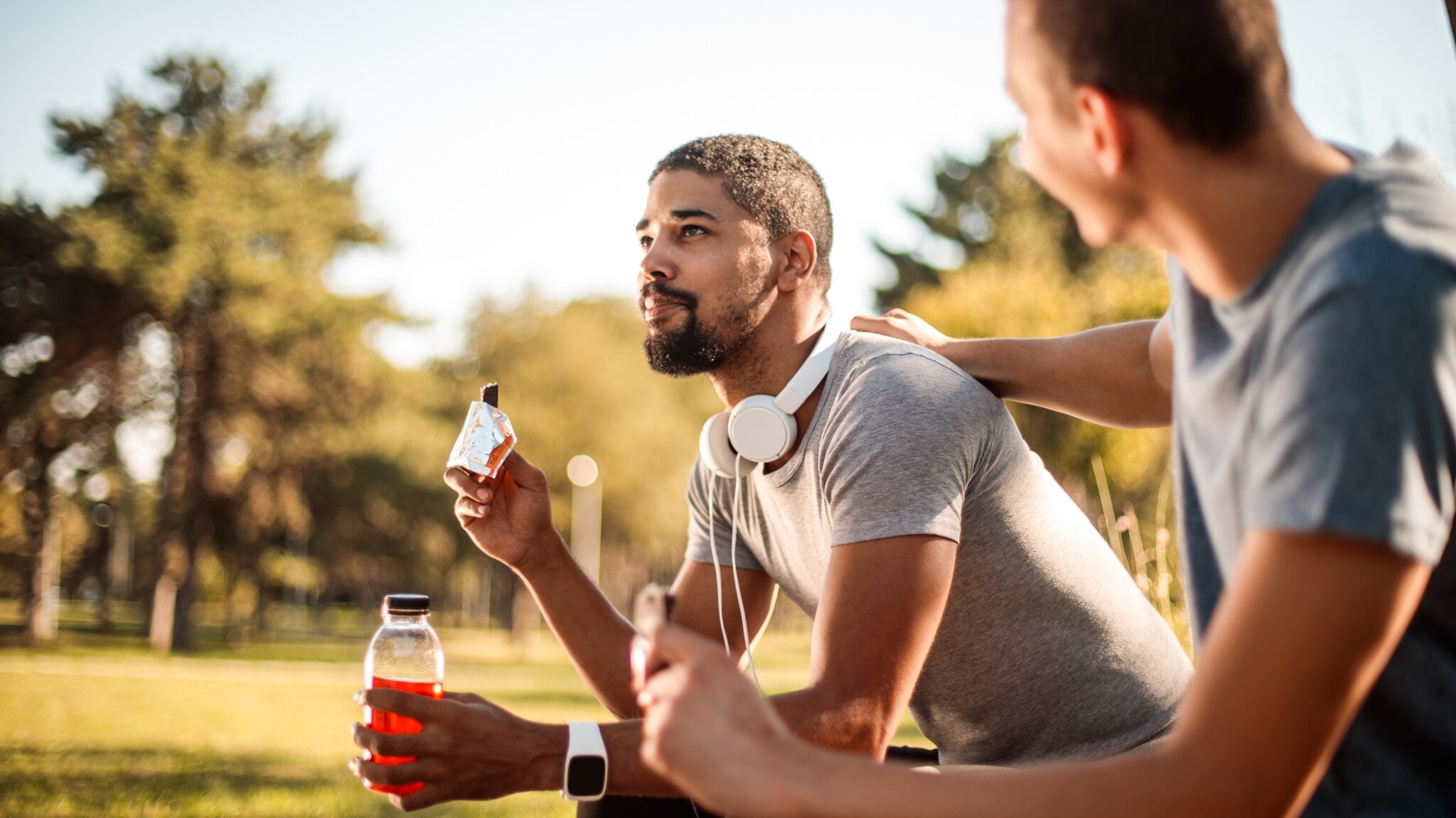 Zwei Männer sitzen draußen auf einer Bank, einer hält eine Wasserflasche und einen Proteinriegel, der andere legt die Hand auf dessen Schulter. Zwei Männer sitzen draußen auf einer Bank, einer hält eine Wasserflasche und einen Proteinriegel, der andere legt die Hand auf dessen Schulter.