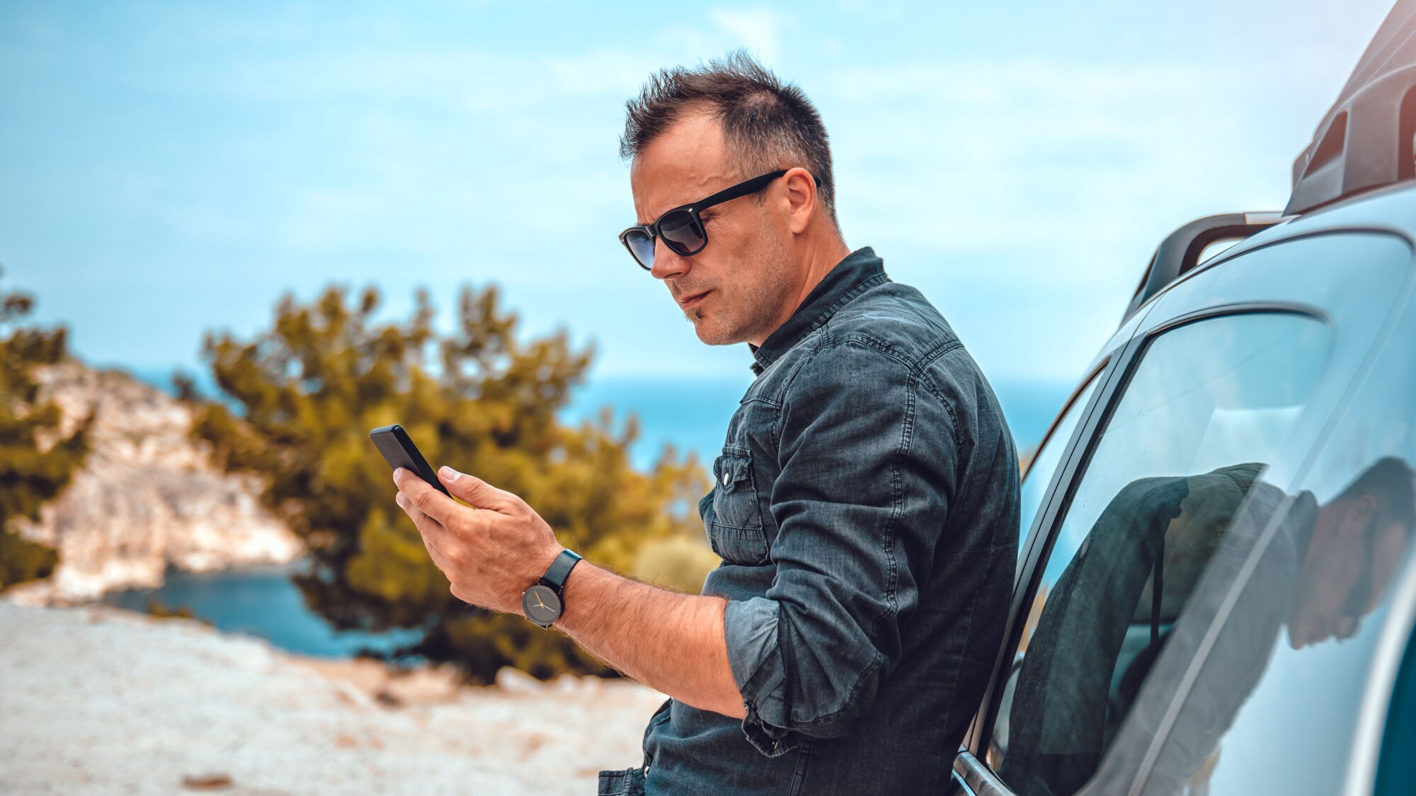 Mann lehnt an silbernem Auto und hält Smartphone in der Hand, trägt dunkles Hemd und Armbanduhr, im Hintergrund Felsen und Bäume unter blauem Himmel.