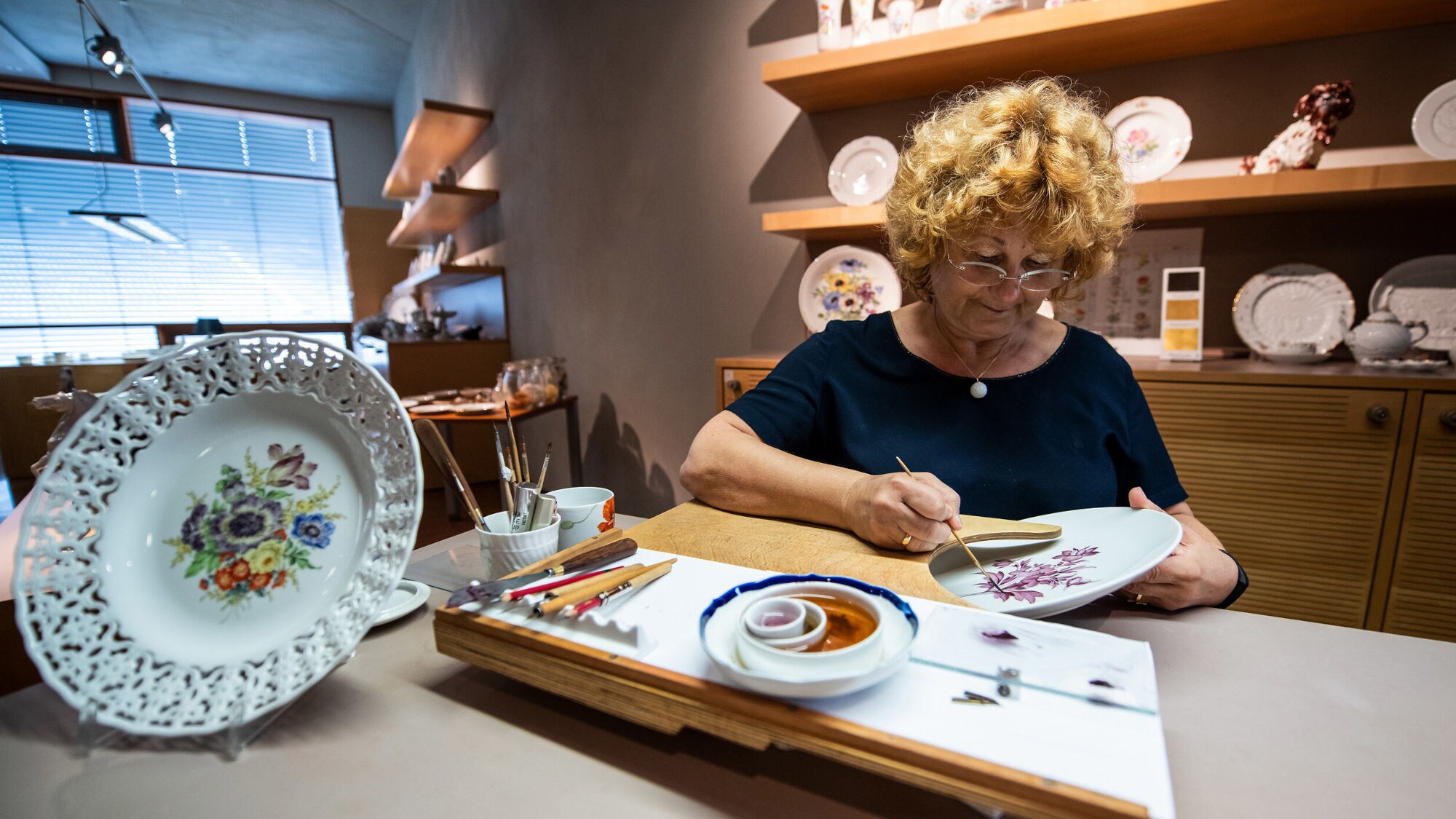 Eine Frau malt mit einem Pinsel ein florales Muster auf einen Porzellanteller in einem Atelier mit ausgestellten Porzellanobjekten. Eine Frau malt mit einem Pinsel ein florales Muster auf einen Porzellanteller in einem Atelier mit ausgestellten Porzellanobjekten.