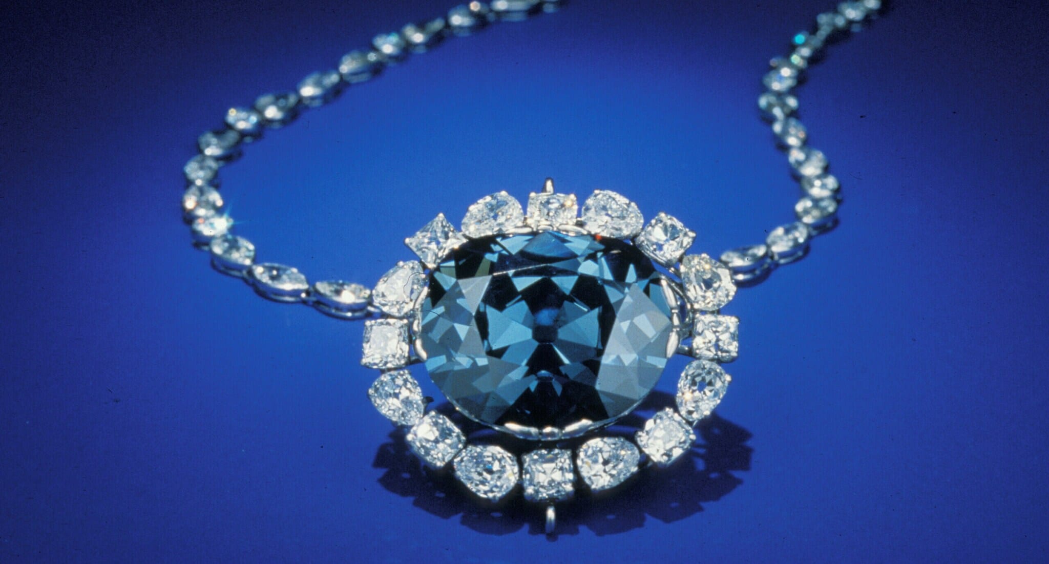 Ein großer, blauer Diamant, eingefasst in kleinere Diamanten an einer Kette auf blauem Untergrund Ein großer, blauer Diamant, eingefasst in kleinere Diamanten an einer Kette auf blauem Untergrund
