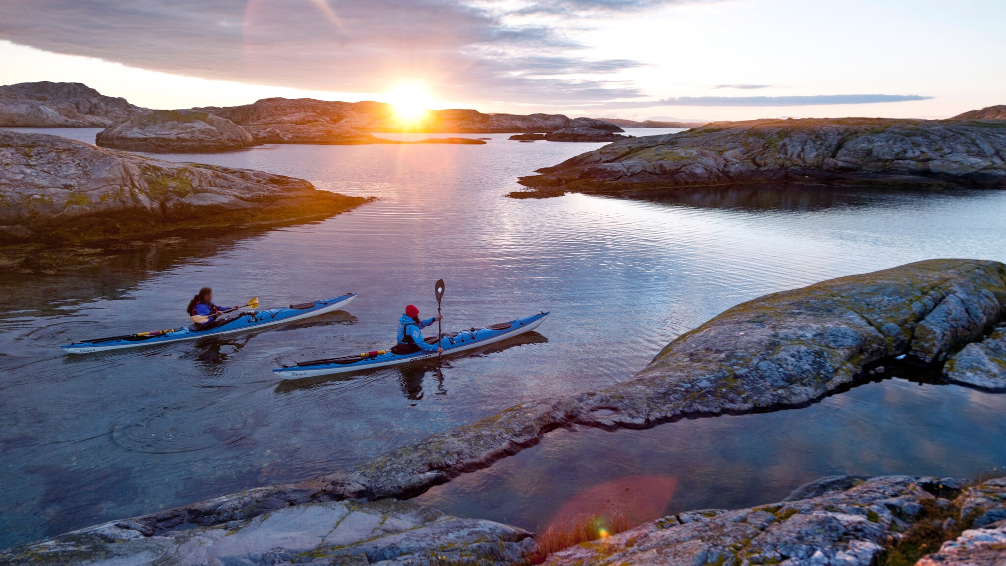 Zwei Personen paddeln in blauen Kajaks auf ruhigem Wasser zwischen felsigen Inseln bei Sonnenuntergang.
