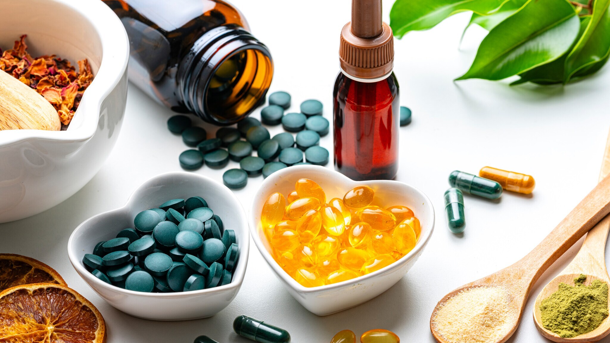 Herzförmige Schalen mit grünen Tabletten und gelben Kapseln, umgeben von weiteren Kapseln, getrockneten Orangenscheiben, zwei Holzlöffeln mit Pulver und grünen Blättern.