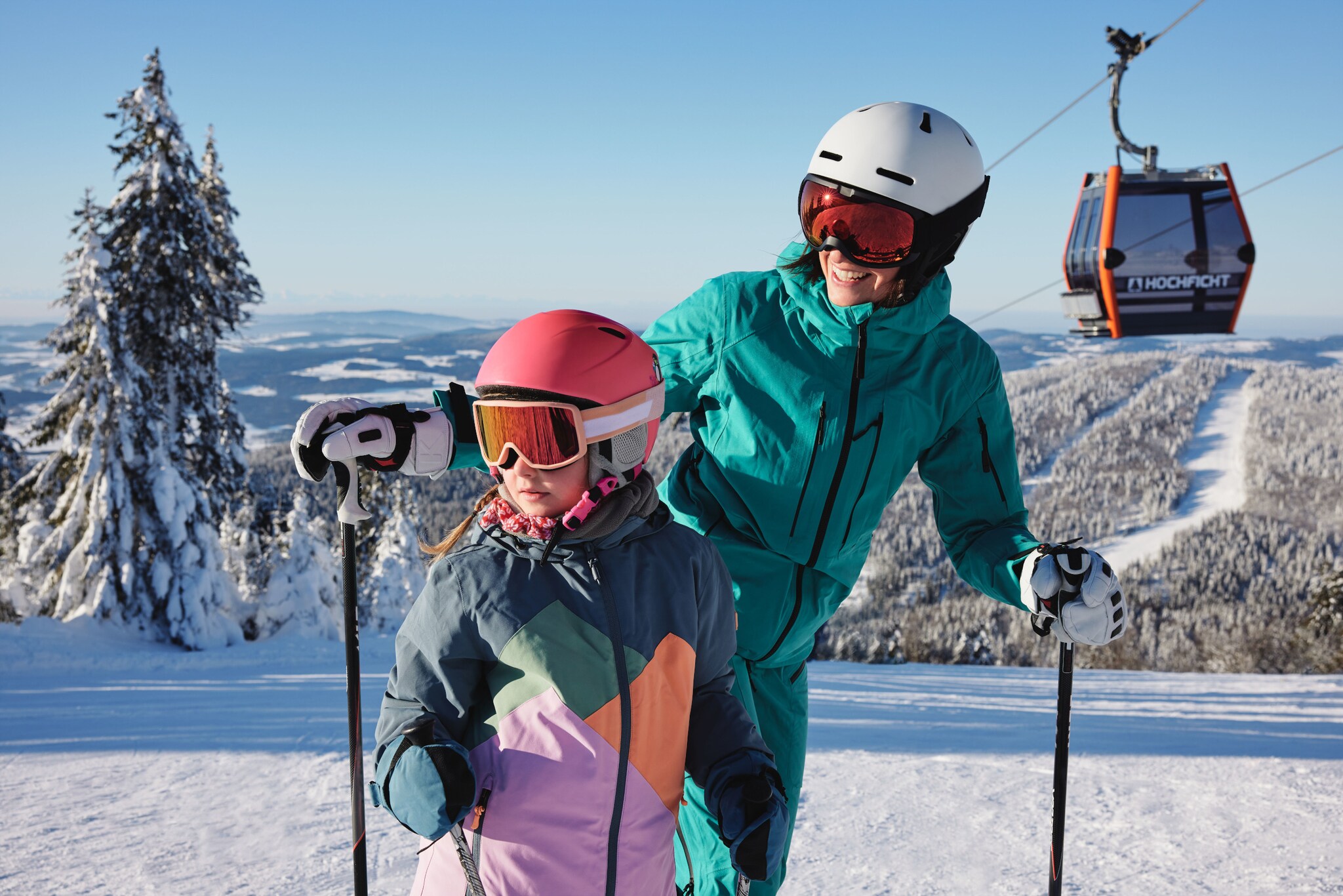 Eine Frau und ein Kind in Skianzügen auf einer Skipiste unter einer Gondel vor verschneiter Waldlandschaft.