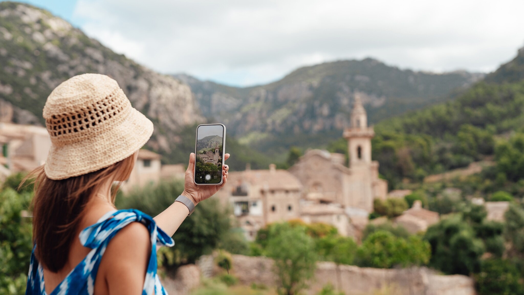 Frau mit Strohhut und blauem Kleid fotografiert Bergdorf mit Smartphone Frau mit Strohhut und blauem Kleid fotografiert Bergdorf mit Smartphone