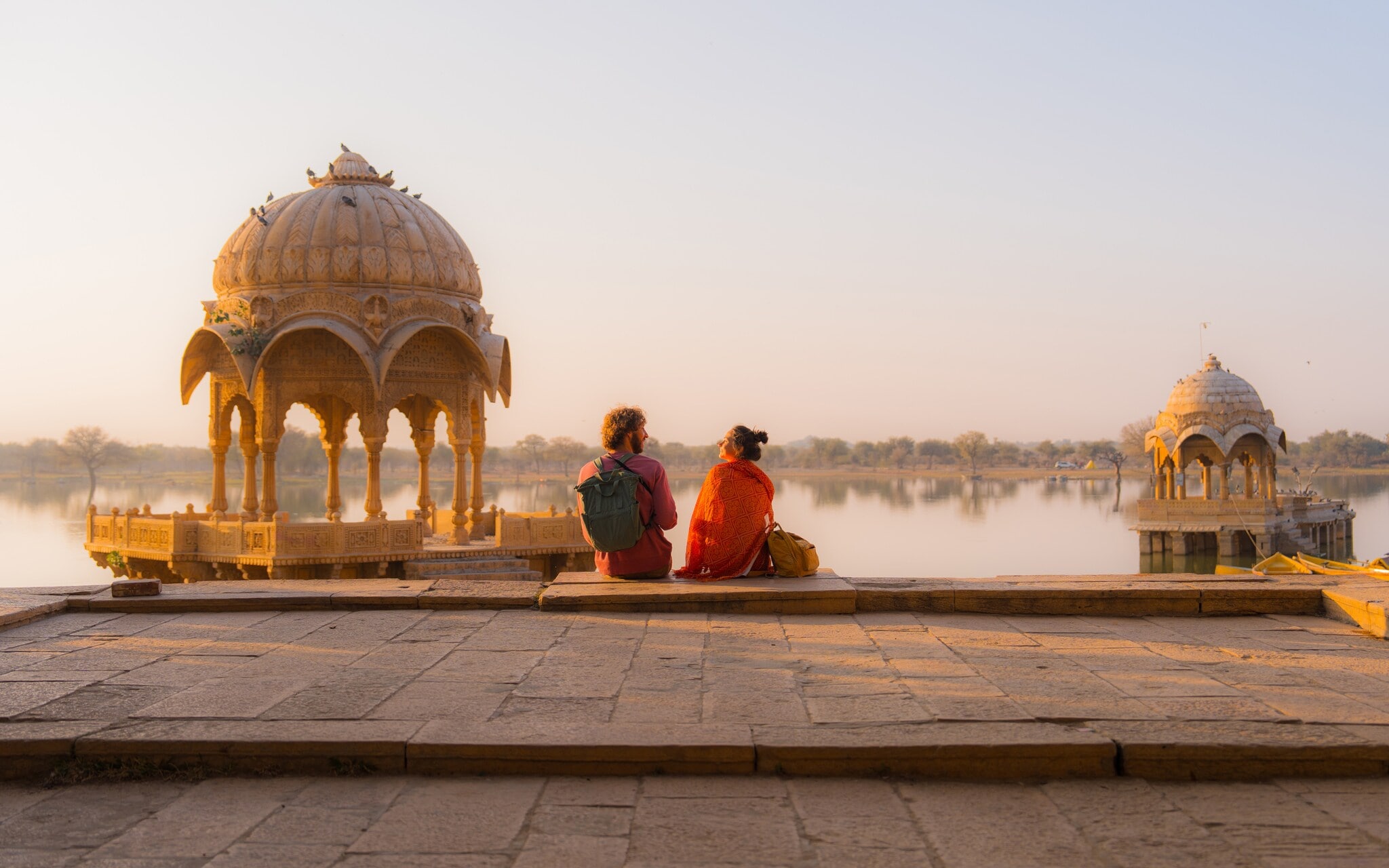 Zwei Personen mit Rucksäcken sitzen in einer buddhistischen Tempelanlage an einem See im weichen Sonnenlicht. Zwei Personen mit Rucksäcken sitzen in einer buddhistischen Tempelanlage an einem See im weichen Sonnenlicht.