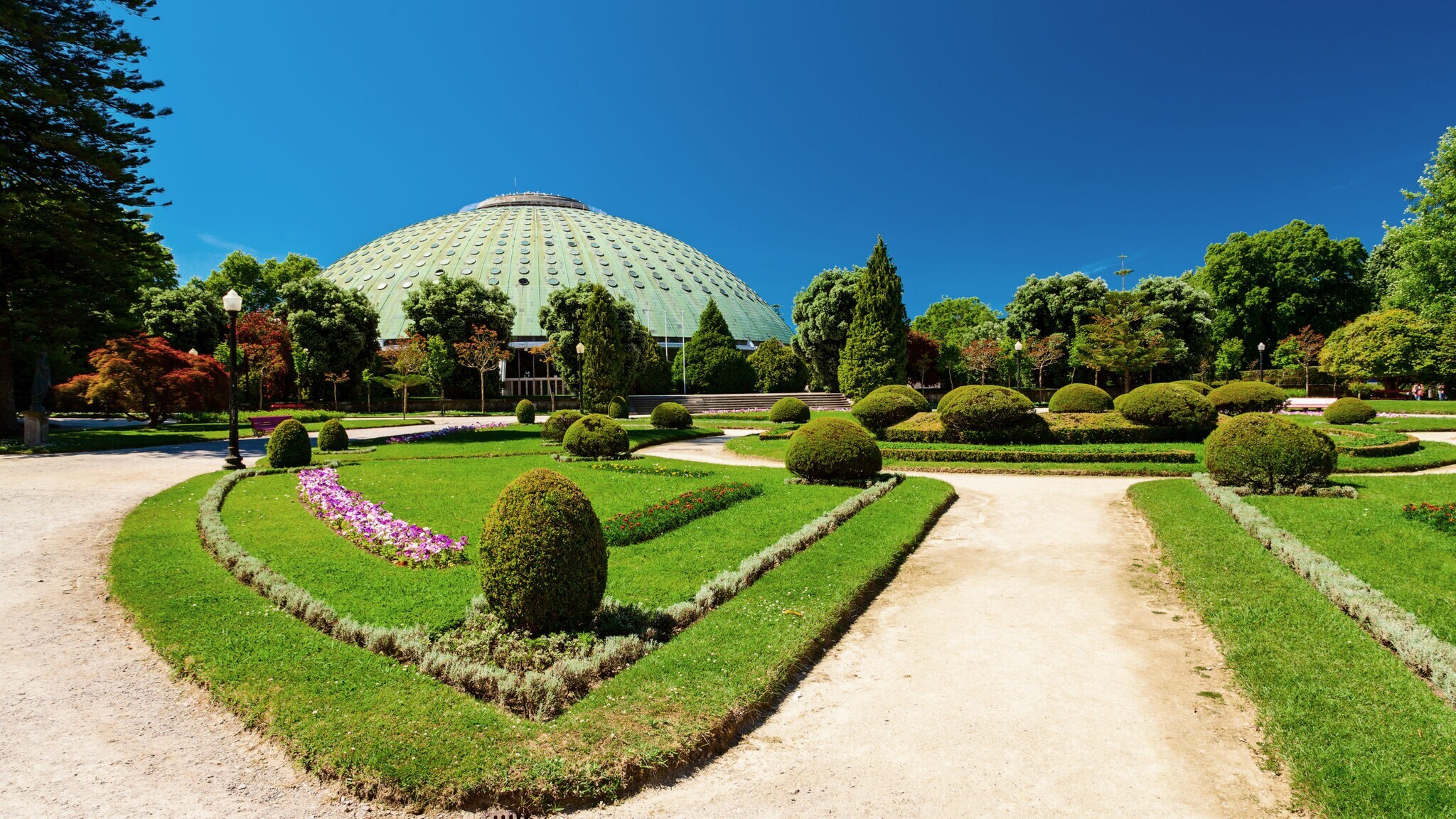 Kuppelgebäude hinter einer gepflegten Parkanlage mit Kieswegen unter blauem Himmel.