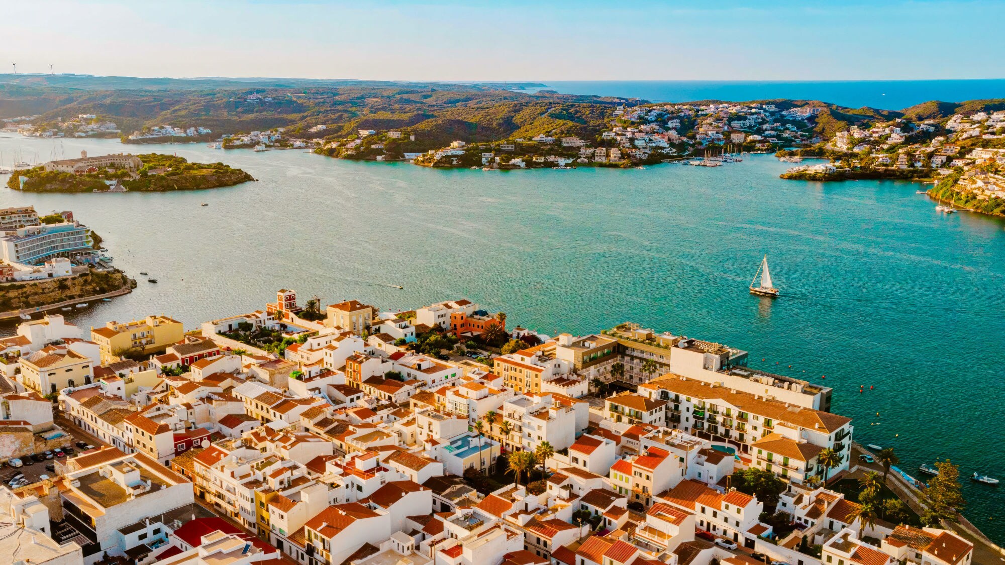 Luftaufnahme einer Küstenstadt mit weißen Gebäuden und roten Dächern, umgeben von Wasser und Hügeln. Ein Segelboot ist auf dem Wasser sichtbar. Luftaufnahme einer Küstenstadt mit weißen Gebäuden und roten Dächern, umgeben von Wasser und Hügeln. Ein Segelboot ist auf dem Wasser sichtbar.